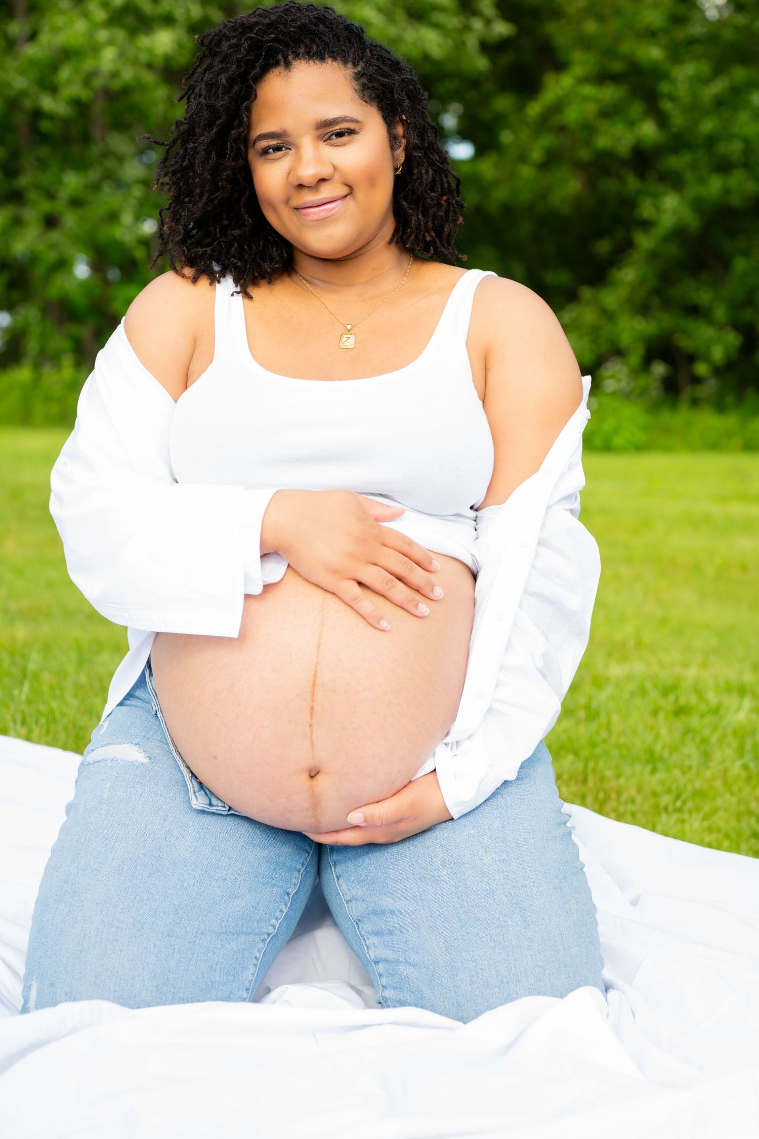 A pregnant woman in a white top and jeans sits outdoors on grass during a maternity photo session.