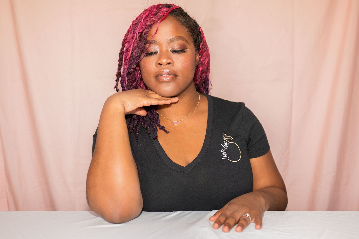 A woman with pink braids wearing a black V-neck top sits at a white table against a pink backdrop.