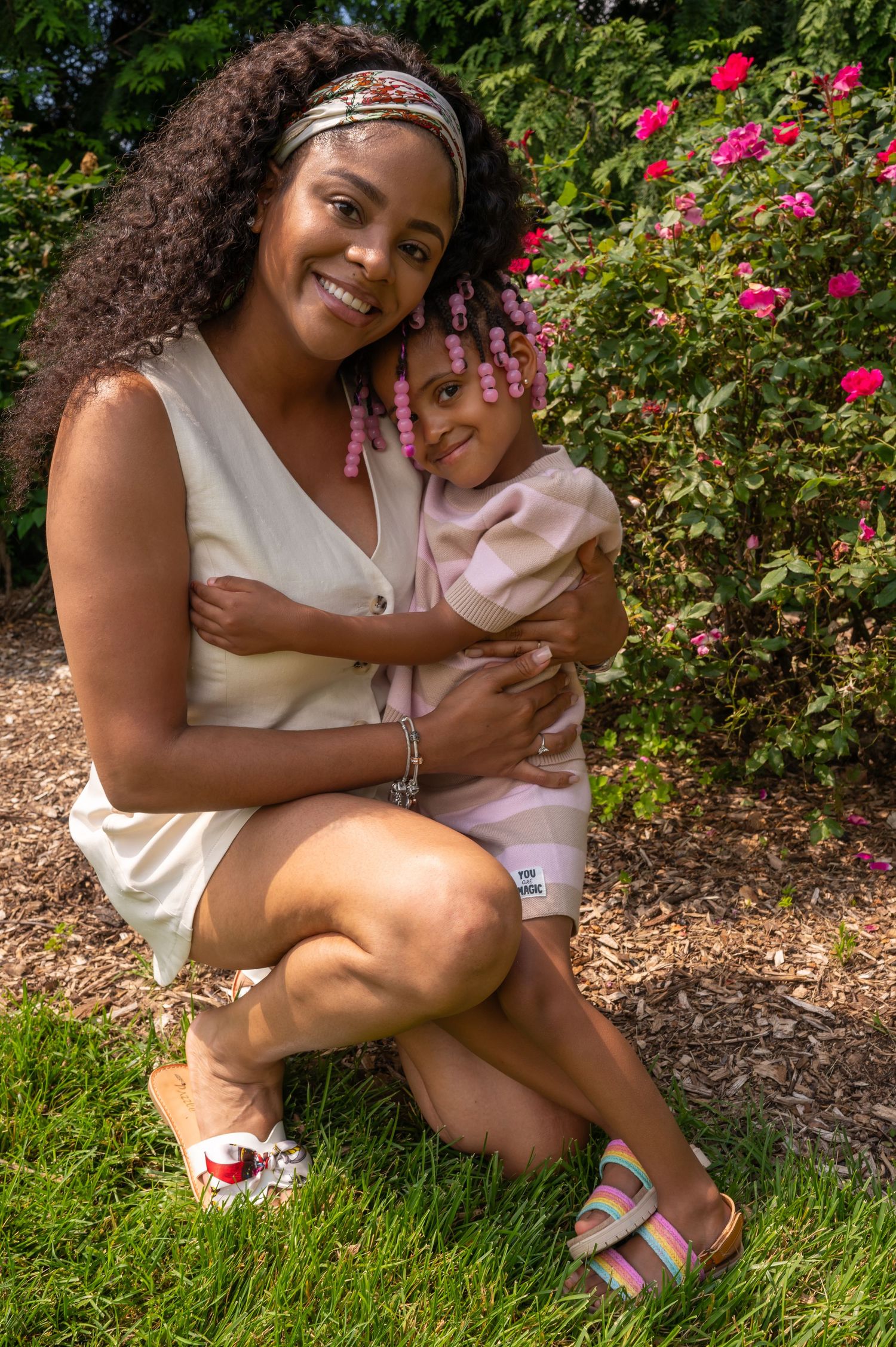 A smiling woman crouches on green grass hugging a young girl with pink braids near a flowering bush outdoors.