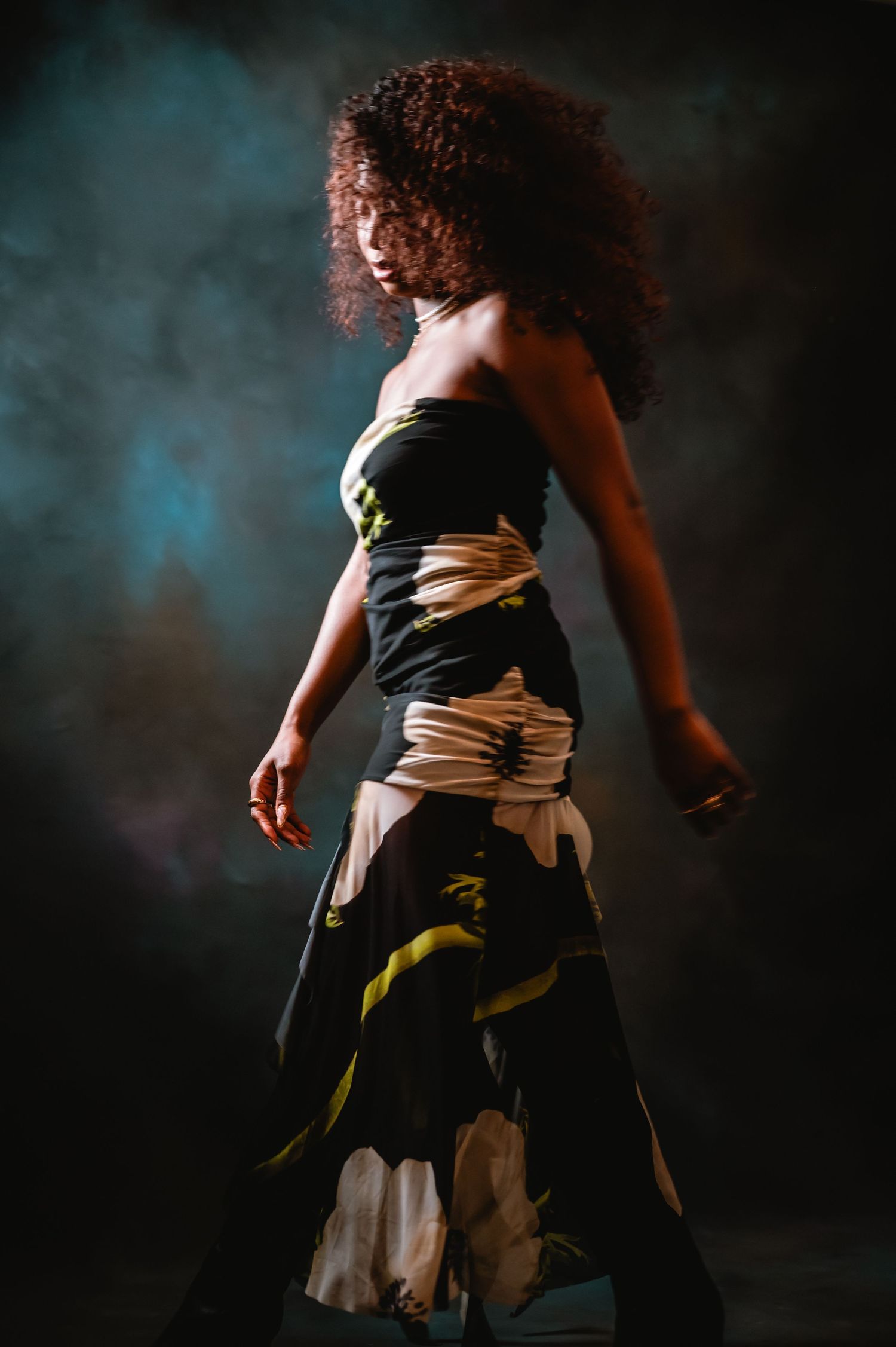 Woman in black floral strapless gown with white sash poses dramatically against dark smoky background.