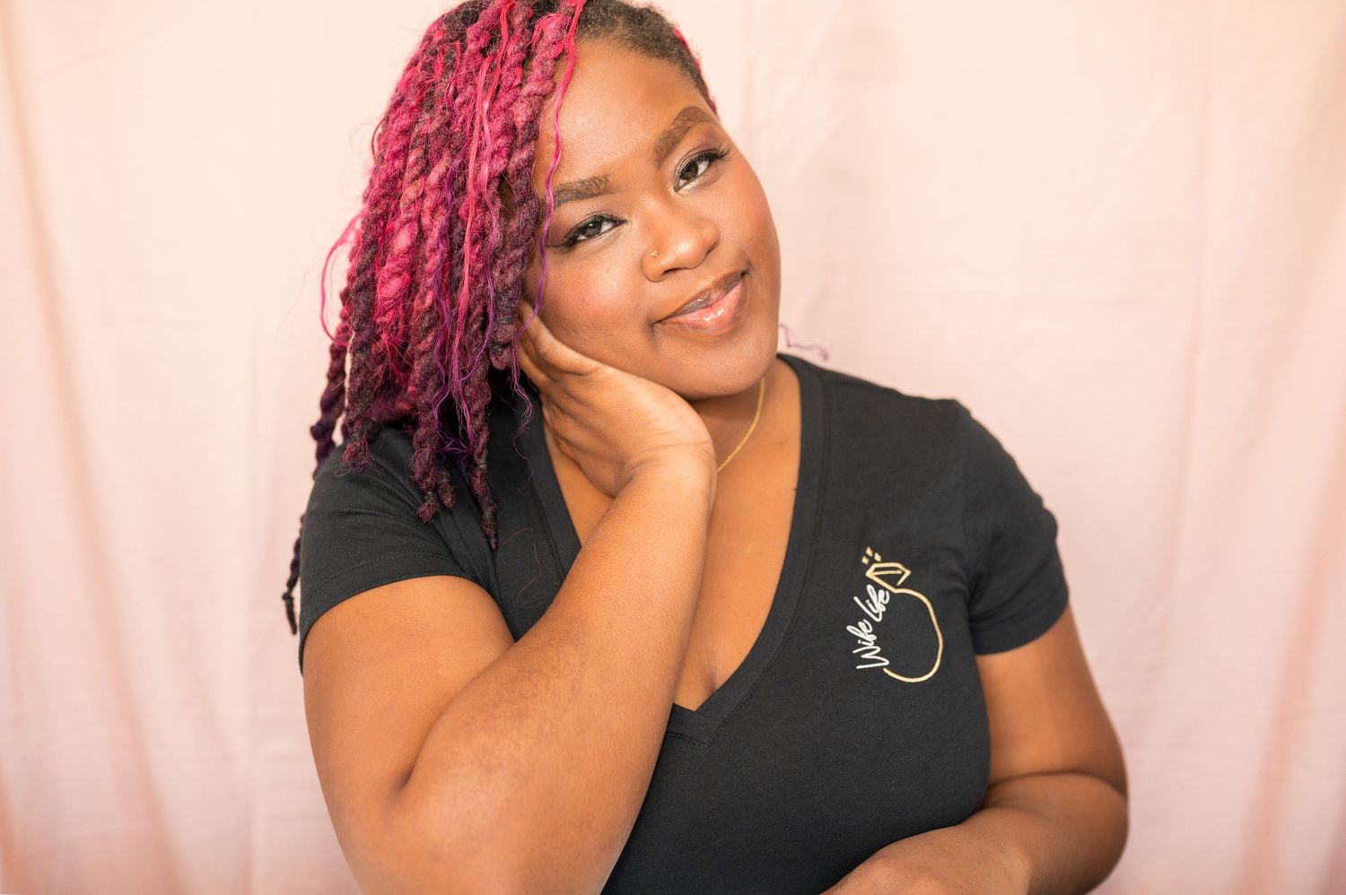 A woman with vibrant pink braids smiles warmly, resting her chin on her hand, wearing a black graphic tee.
