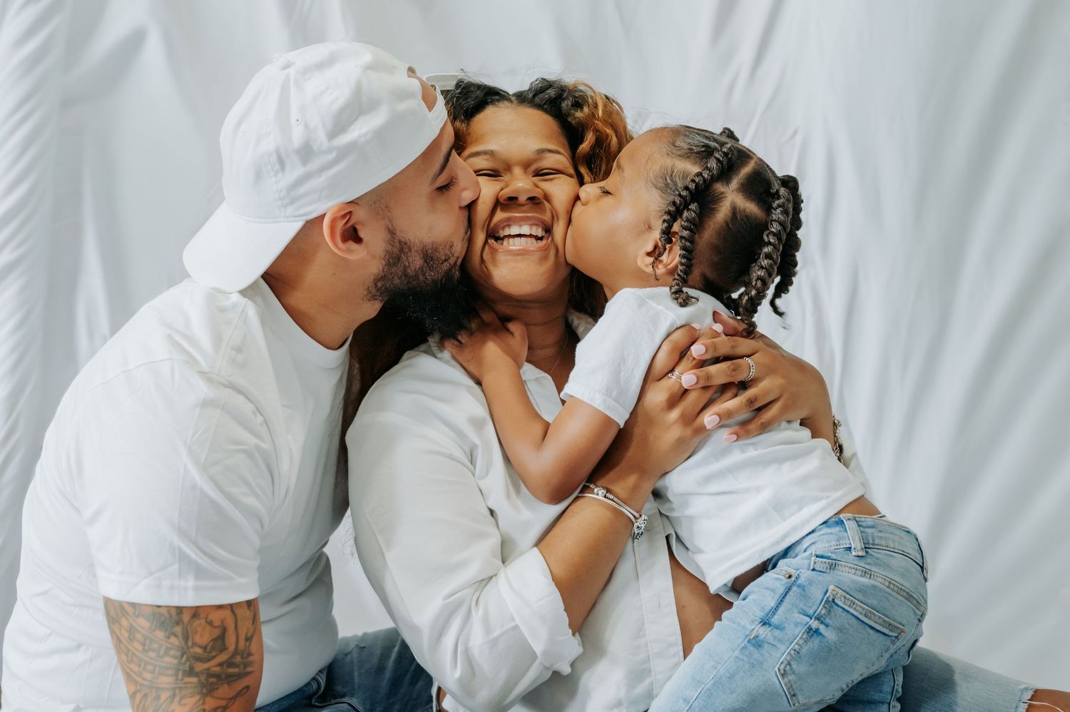 Happy family moment with parents kissing their smiling daughter on cheeks, all wearing white outfits.