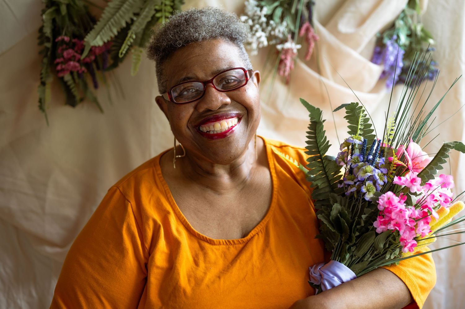 Smiling woman in orange top holding colorful pink and blue flowers, wearing red glasses, posing indoors.