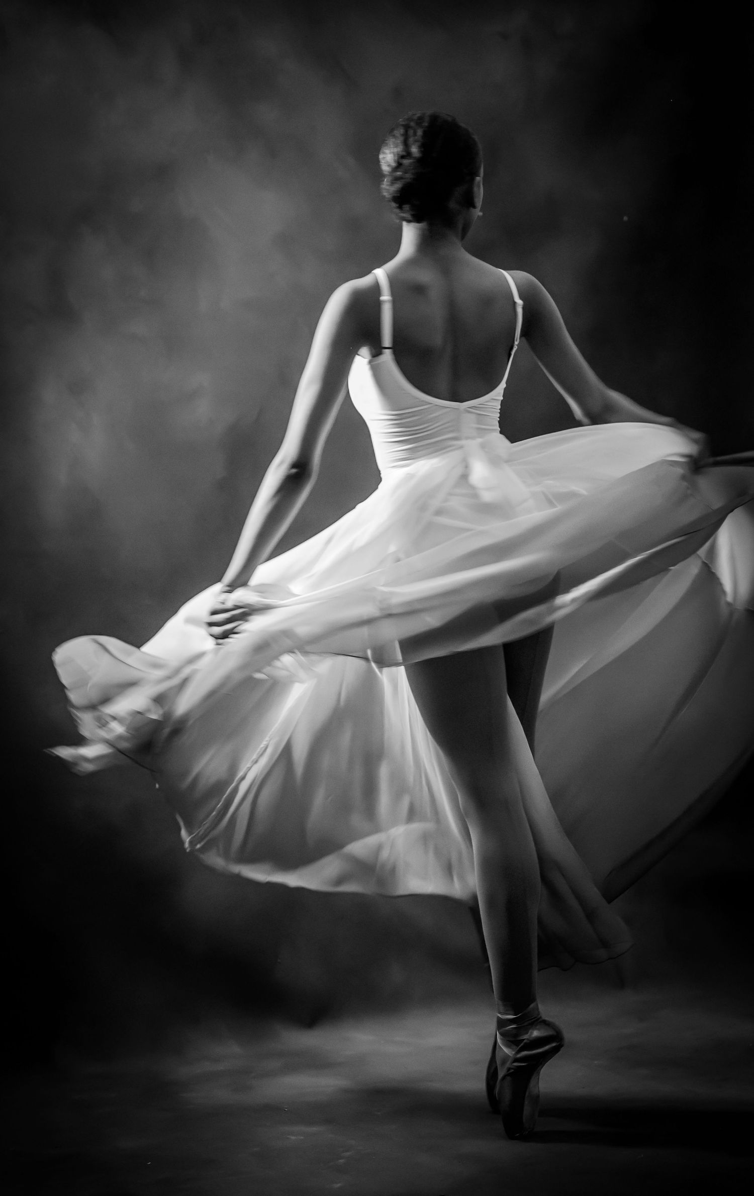Black and white photo of a graceful ballerina in a flowing white tutu dancing en pointe, viewed from behind.