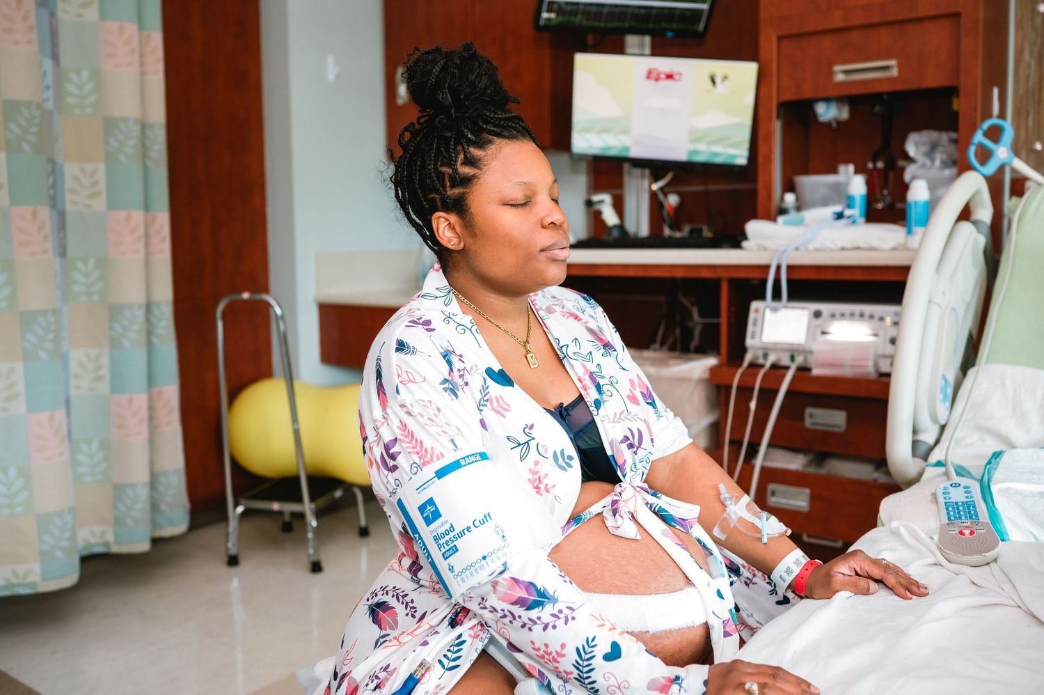Patient in floral hospital gown rests in medical room chair during treatment with IV equipment nearby.