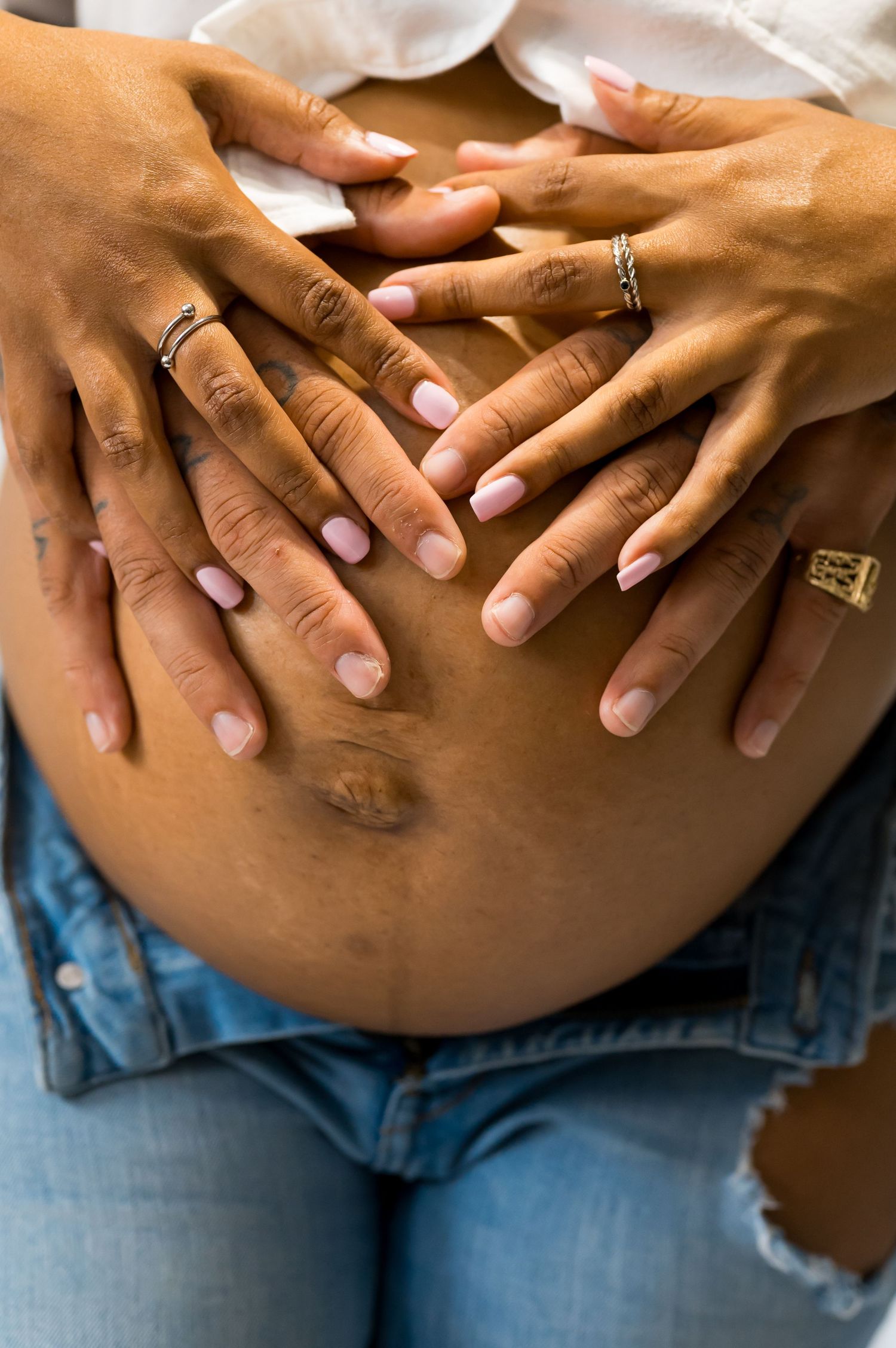 Two pairs of hands cradling a pregnant belly, wearing rings, with manicured pink nails over denim jeans.