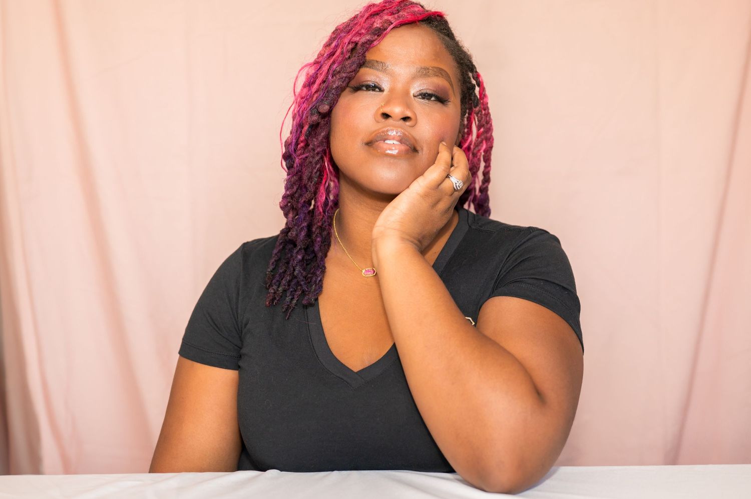 A woman with pink braided hair poses thoughtfully with her hand on her cheek against a soft pink background.