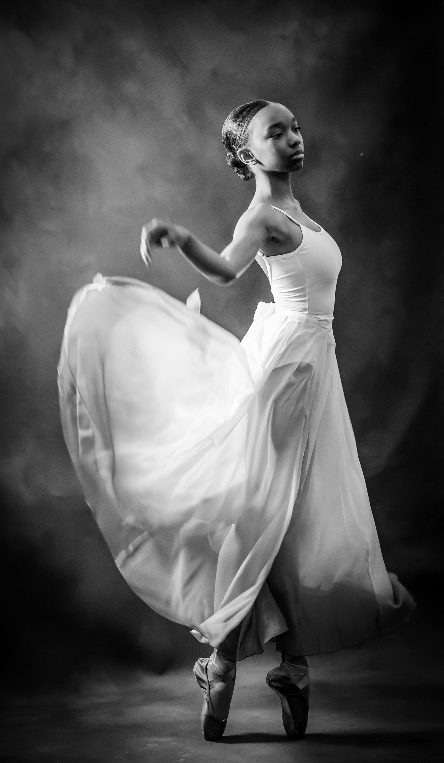 Black and white photo of a graceful ballerina in white dress on pointe, performing an elegant dance pose.