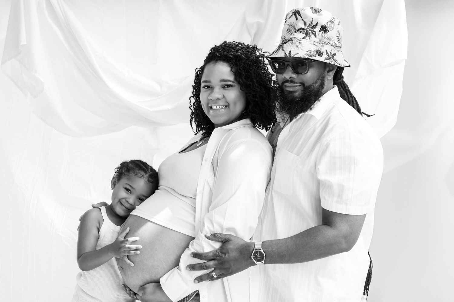 A loving family poses together in a white studio setting with a child and expectant mother in black and white photography.