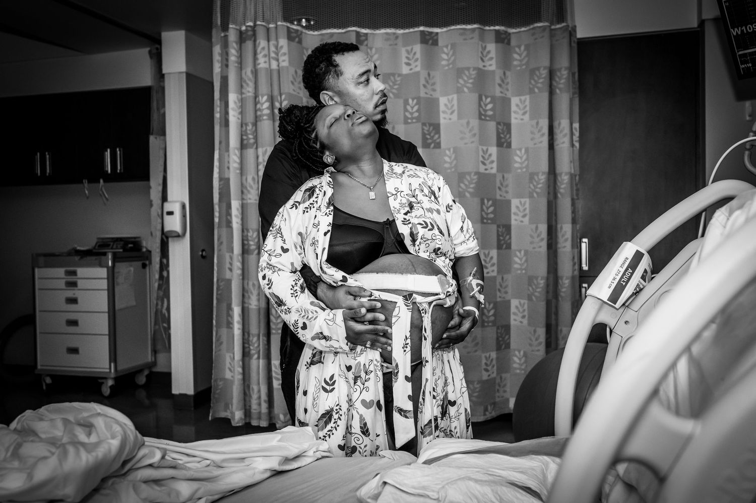A person wearing a patterned robe stands in a medical room beside a hospital bed in black and white.