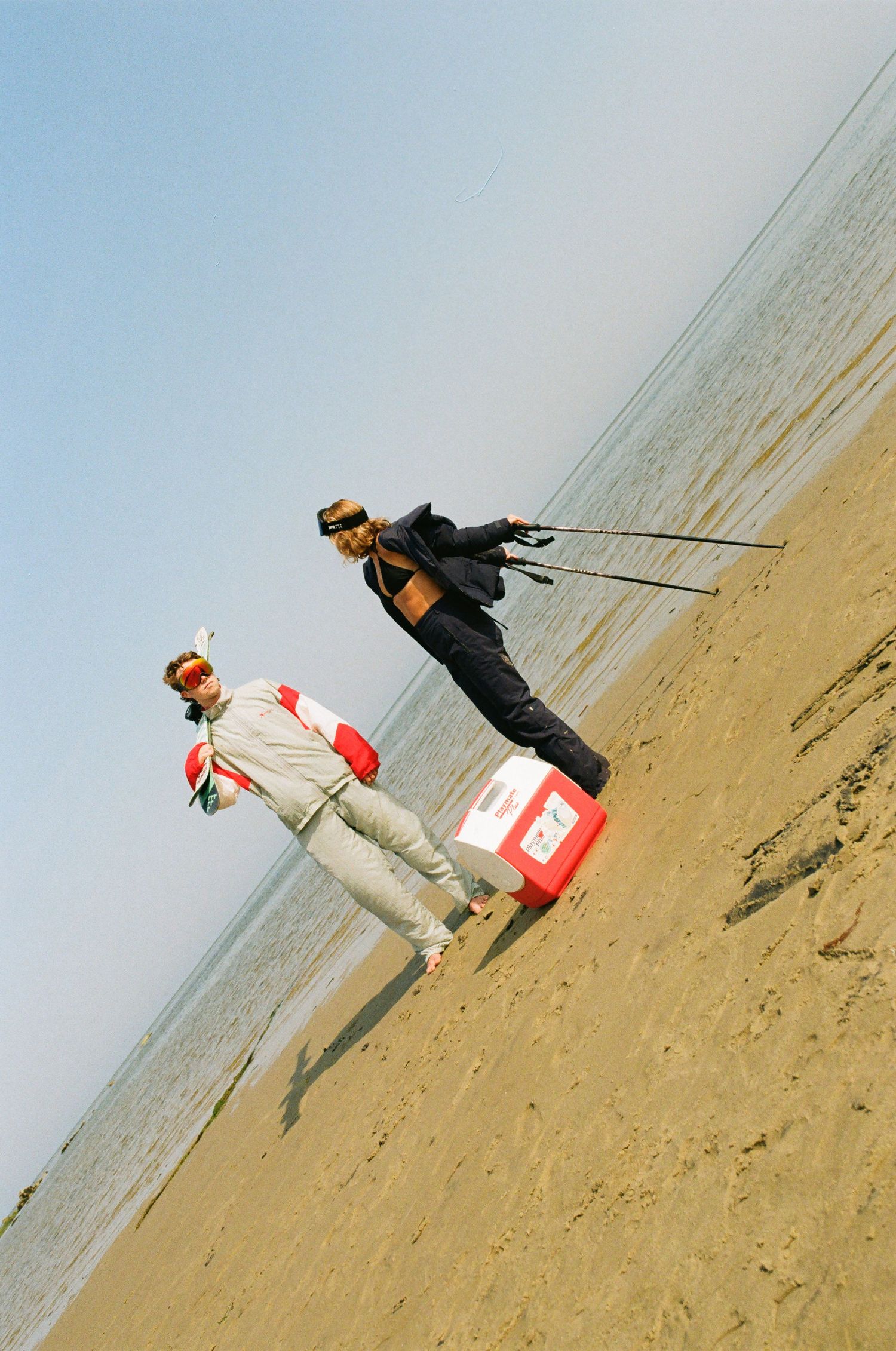Two men pose on a sandy beach, one standing on a red cooler holding kite strings, under a clear blue sky.