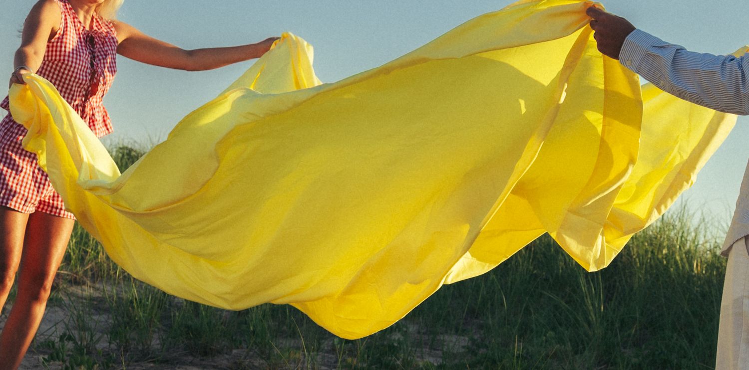 Two people holding a flowing yellow fabric billowing in the wind outdoors on a sunny day.