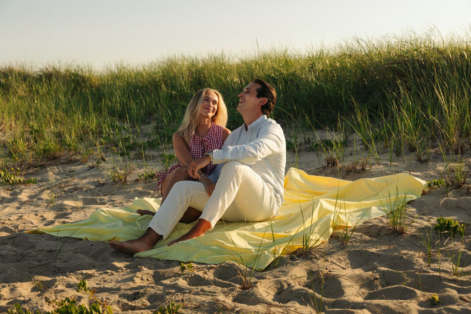 Couple sitting on yellow blanket on sandy beach near grass dunes at sunset.