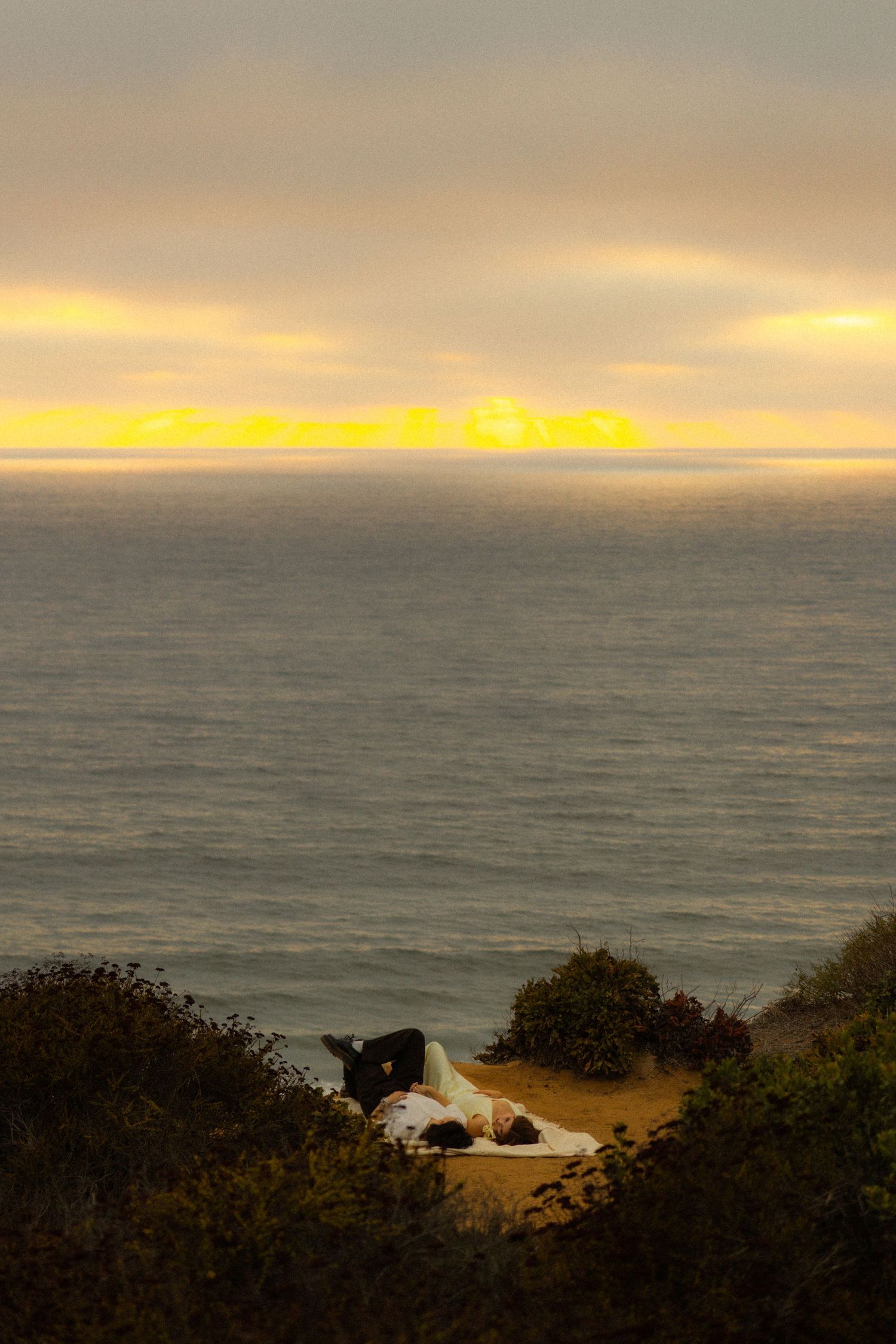 A tent sits on a coastal cliff overlooking the ocean at sunset with golden clouds illuminating the horizon.