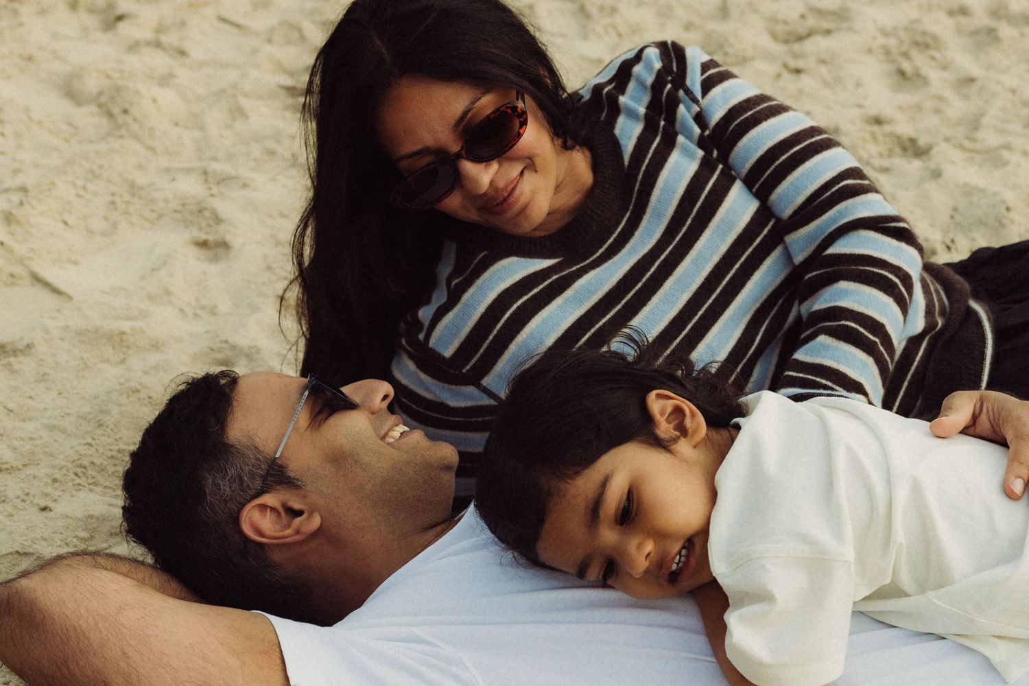 Family of three relaxing on sandy beach, parents lovingly gazing at sleeping baby in white outfit.