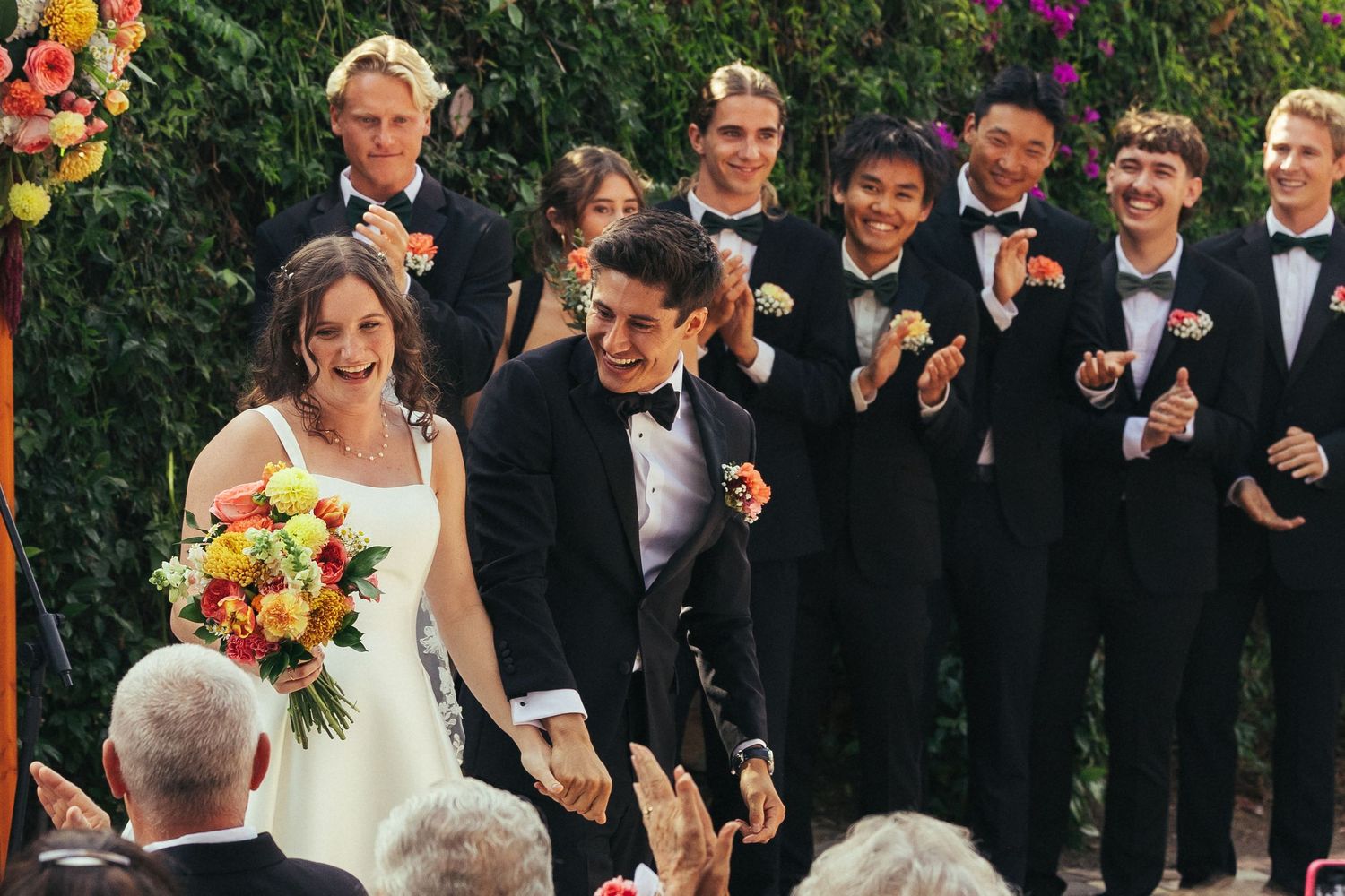 Happy couple walks down the aisle at outdoor wedding ceremony with groomsmen in black tuxedos celebrating behind them.