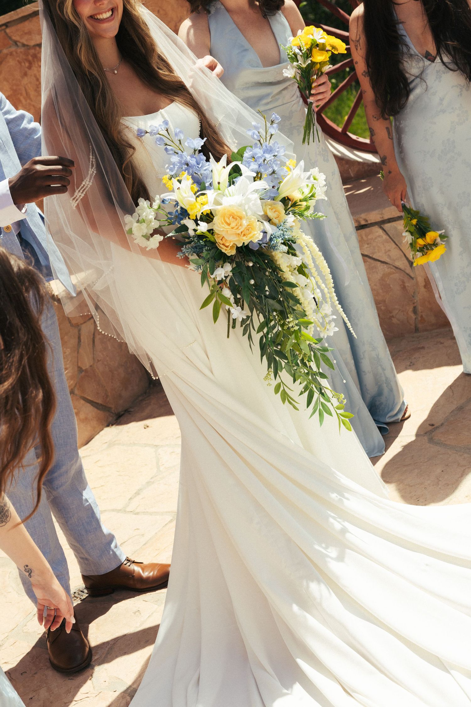 Bride in white gown holding cascading bouquet of yellow roses and blue flowers surrounded by wedding party in sunlight.