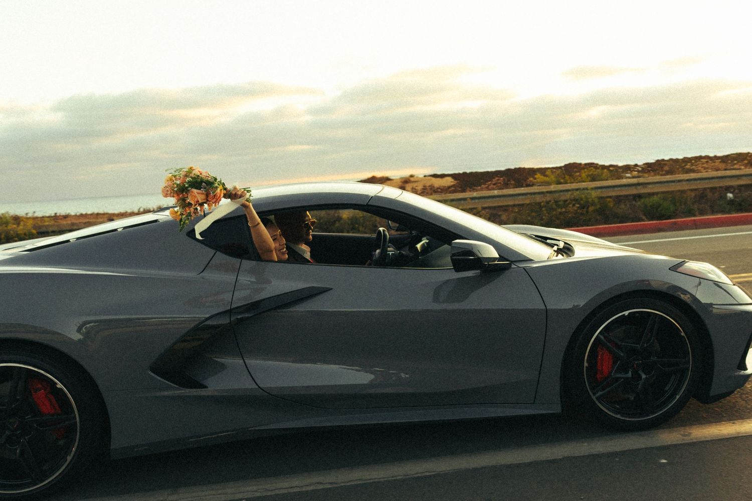 Bride with flower crown riding in a gray Chevrolet Corvette C8 sports car on a scenic highway at sunset.