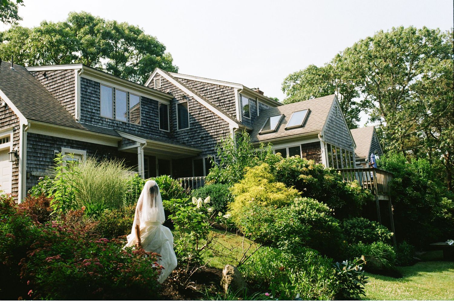 Bride in white veil standing in lush garden beside charming Cape Cod shingle-style home surrounded by greenery.