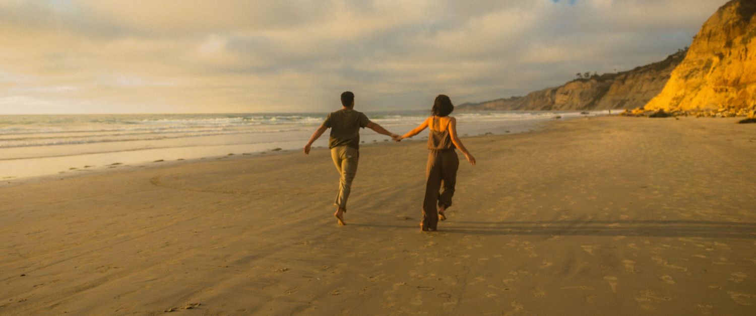 Two people run together along a scenic beach at sunset with golden cliffs in the background.