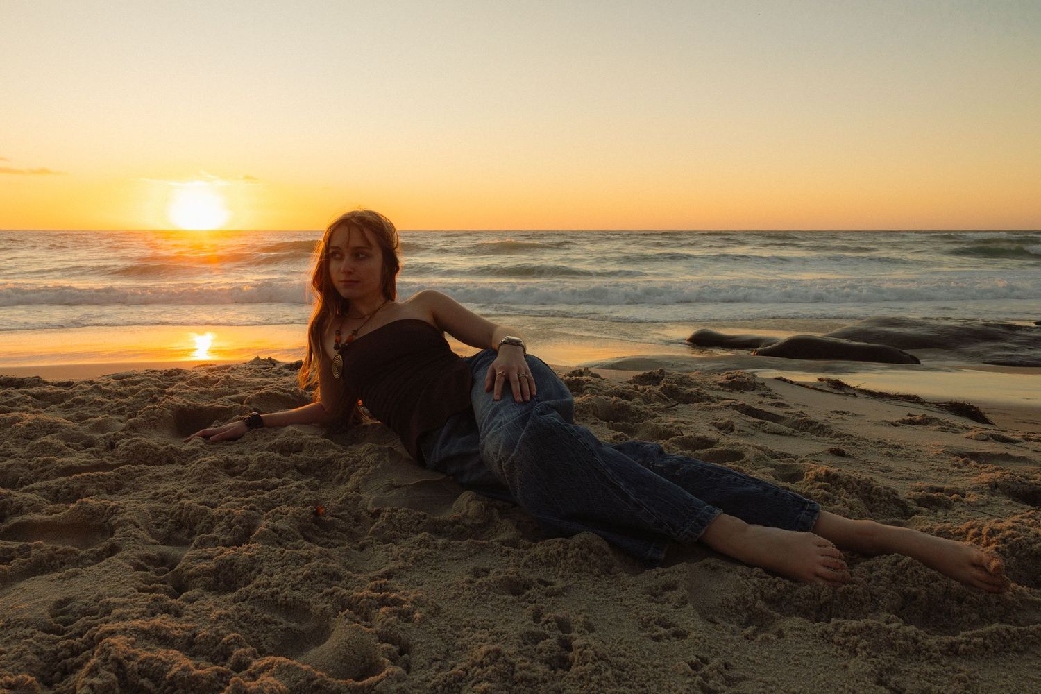 A person lies on a sandy beach at sunset as golden light illuminates the ocean waves and horizon.