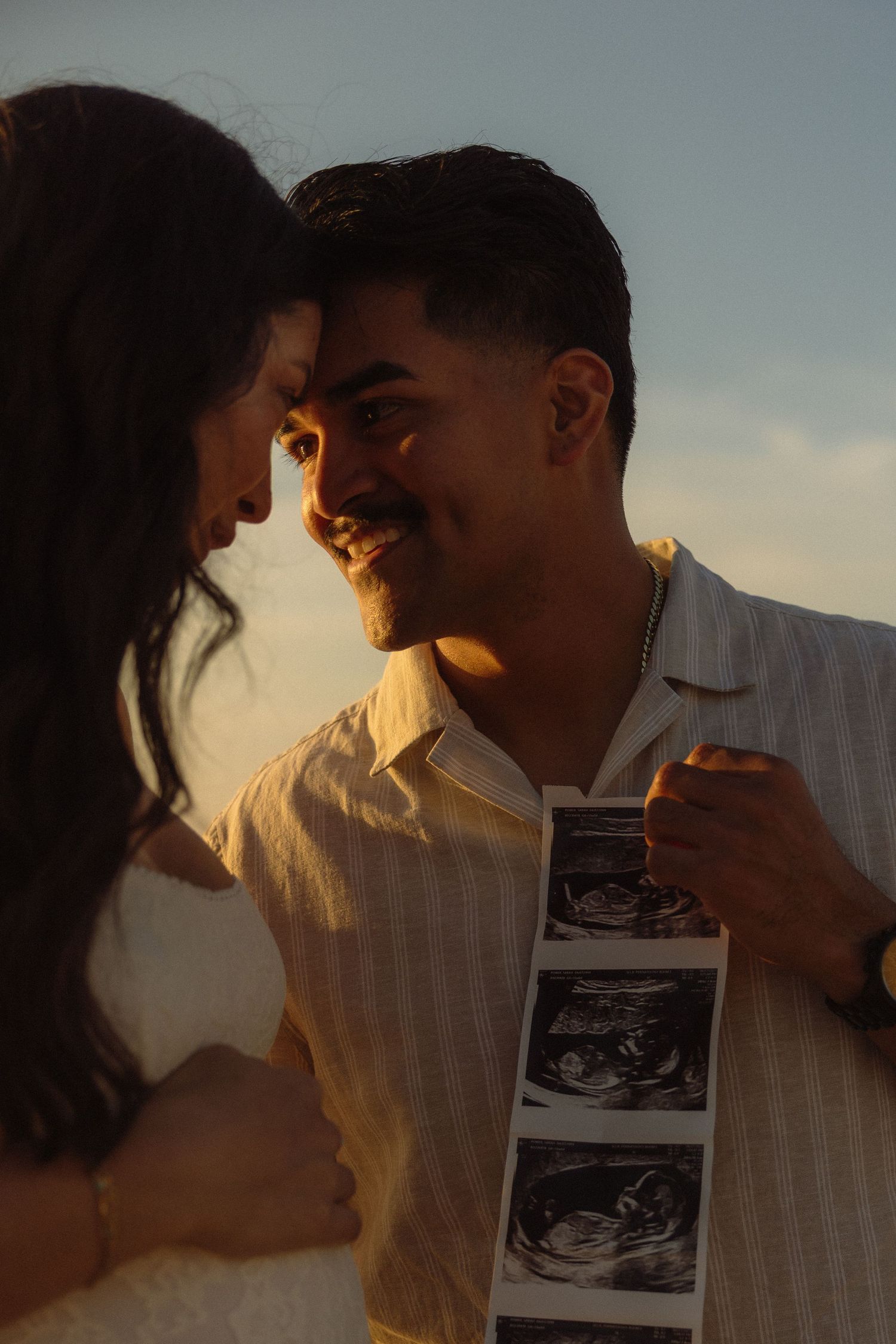 A couple shares an intimate moment at sunset while holding ultrasound photos in a pregnancy announcement scene.