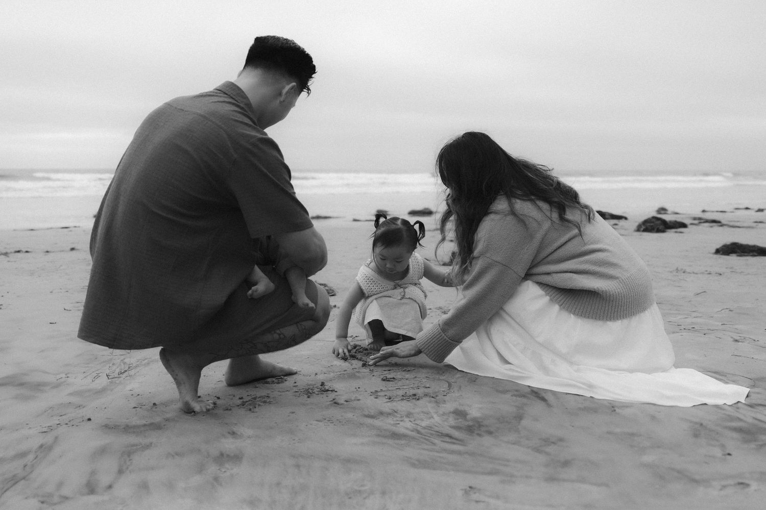 Two people taking pictures on a sandy beach during sunset while kneeling down in black and white.