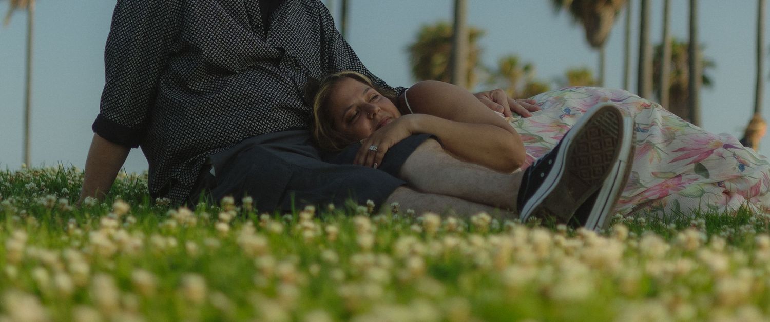 A person relaxes lying down on grass scattered with daisies as palm trees sway in the background.