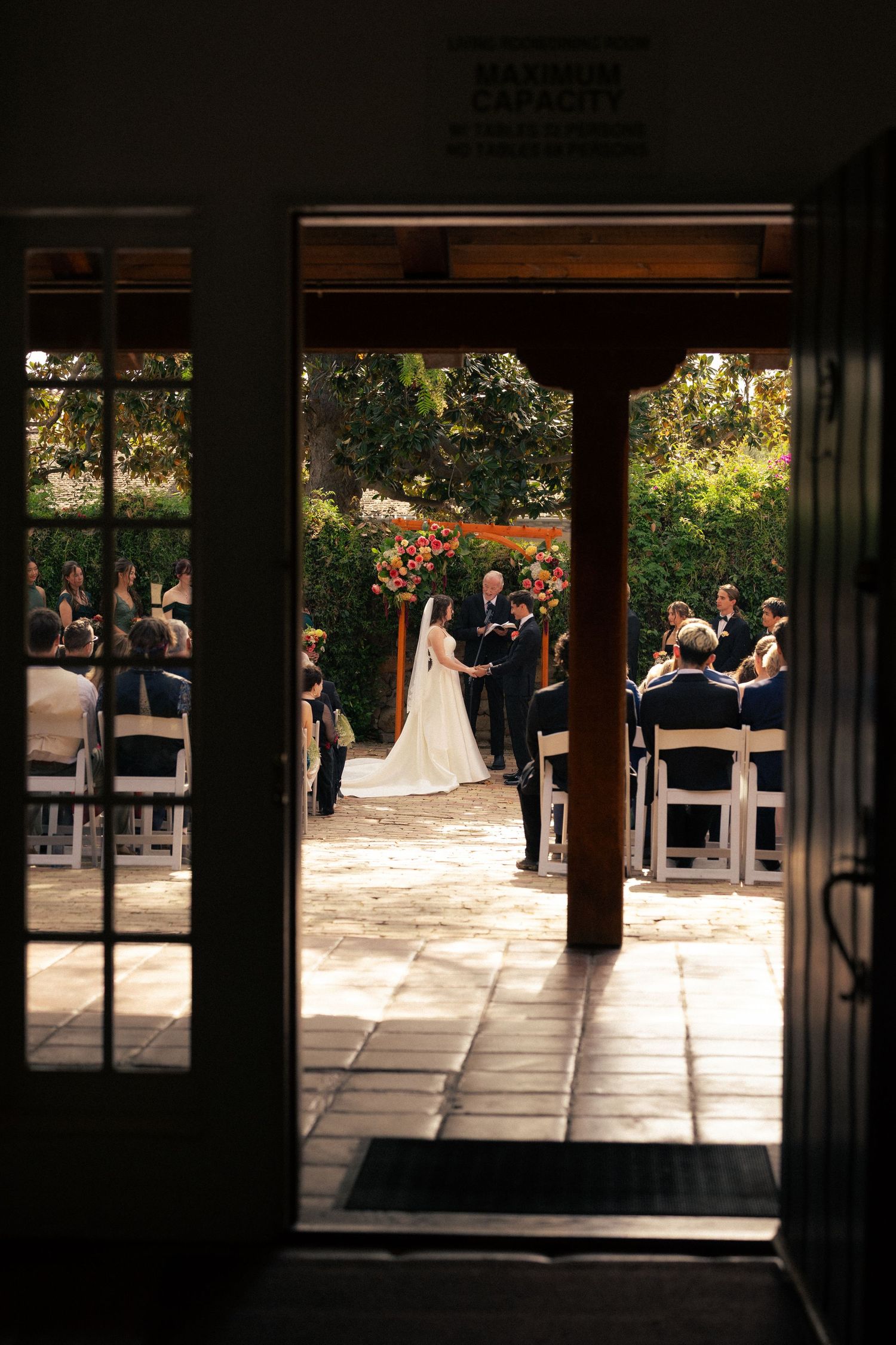 Outdoor wedding ceremony viewed through open doors, bride and groom at altar with guests seated on white chairs.