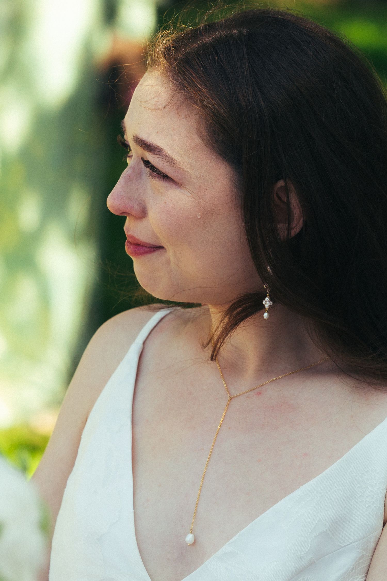 A bride in a white dress wearing pearl drop earrings and a delicate gold necklace, gazing softly to the side outdoors.