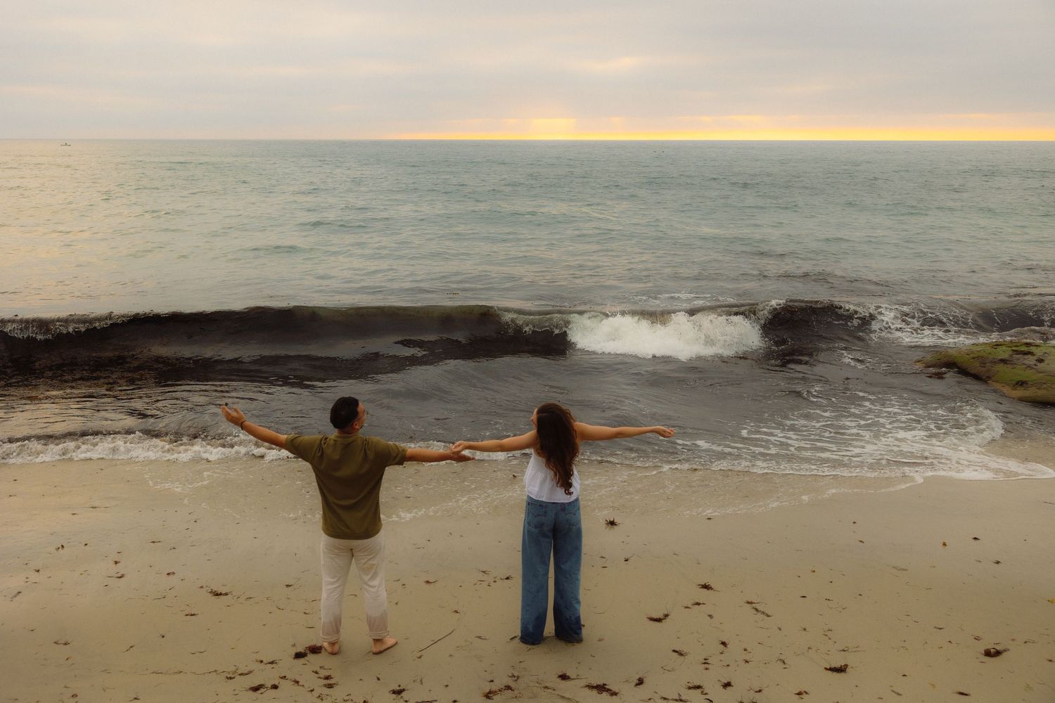 Two people stand with outstretched arms on a beach at sunset as waves crash against rocks on the shoreline.