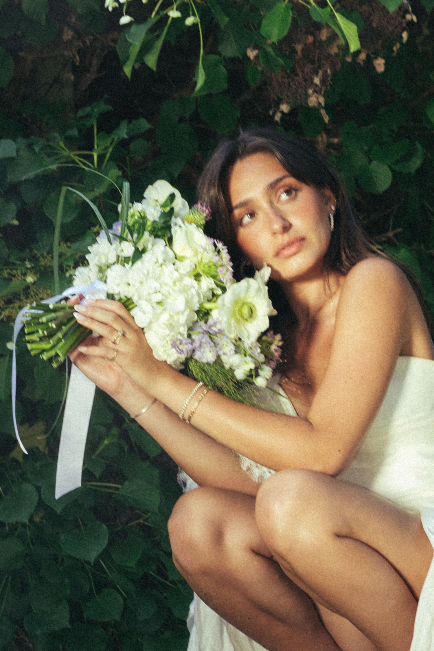Bride crouching in white dress holds white floral bouquet with ribbon against green garden backdrop.