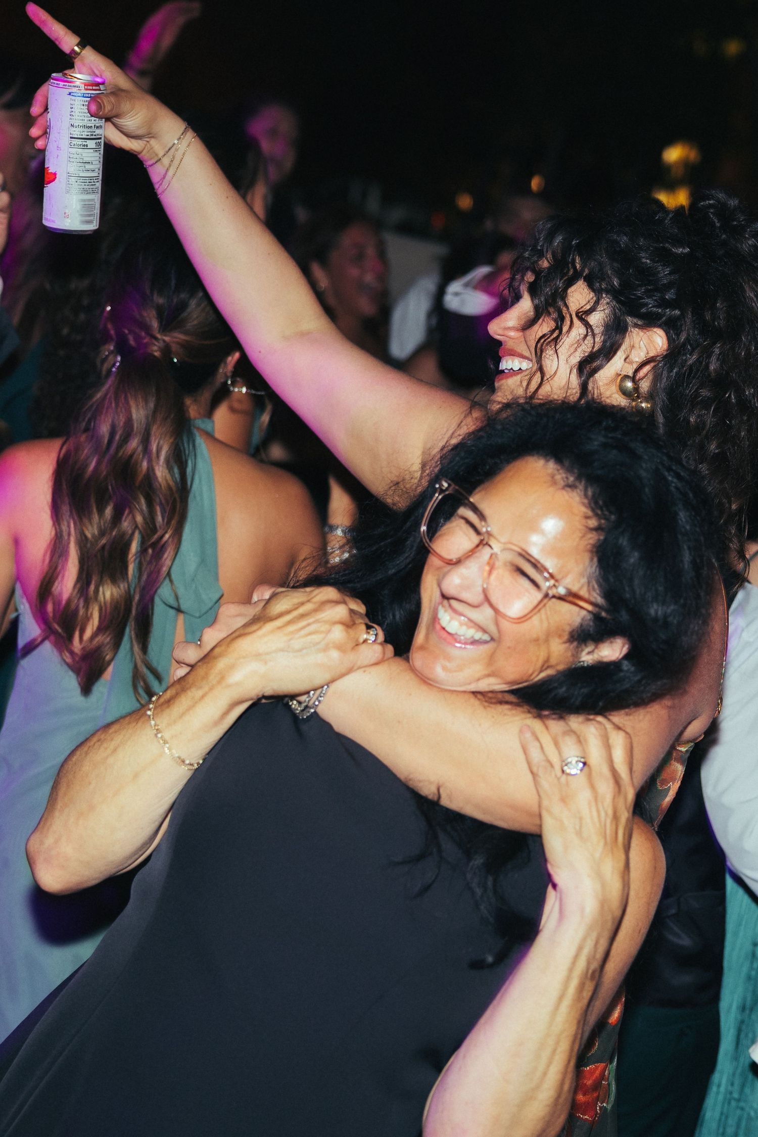 Two women dancing and embracing joyfully at a lively party, one holding a drink can in the air.