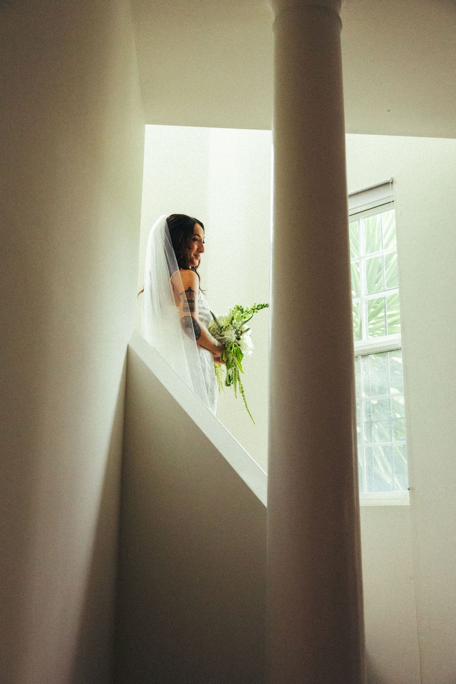 Bride in white gown and veil holding green bouquet stands near window on staircase landing in elegant venue.