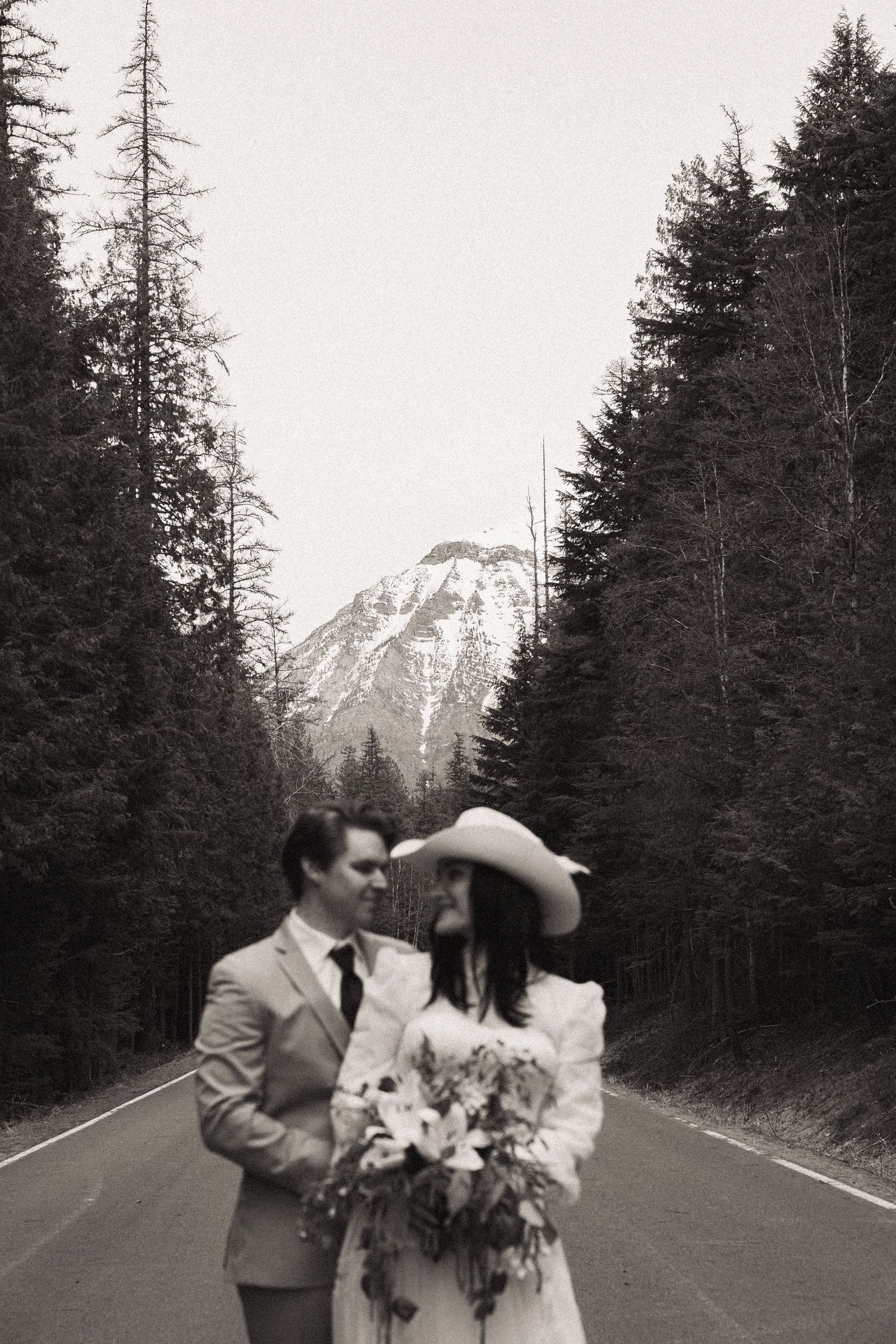 Black and white photo of a couple in wedding attire on a forest road with a snow-capped mountain backdrop.