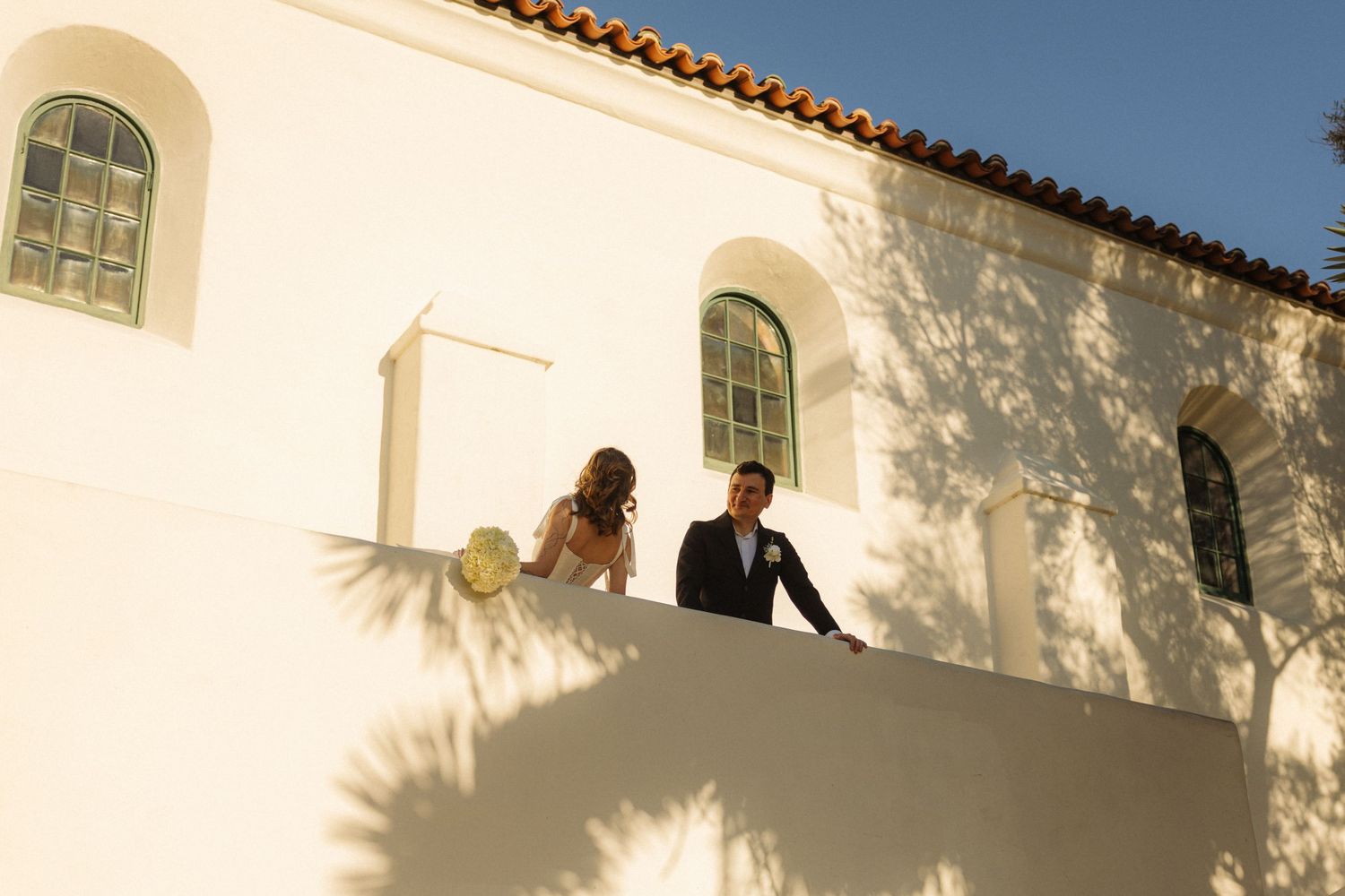 A couple stands on a white Spanish-style balcony during golden hour as shadows dance across the stucco walls.