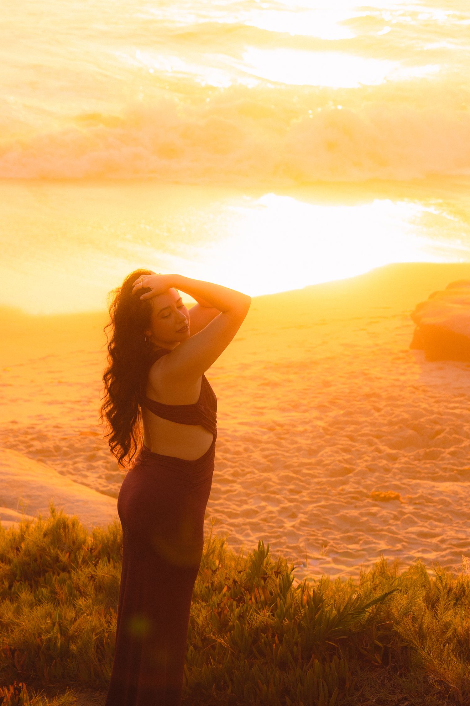 Silhouette of a person with flowing hair stands on a cliff edge against a vibrant orange and yellow sunset sky.
