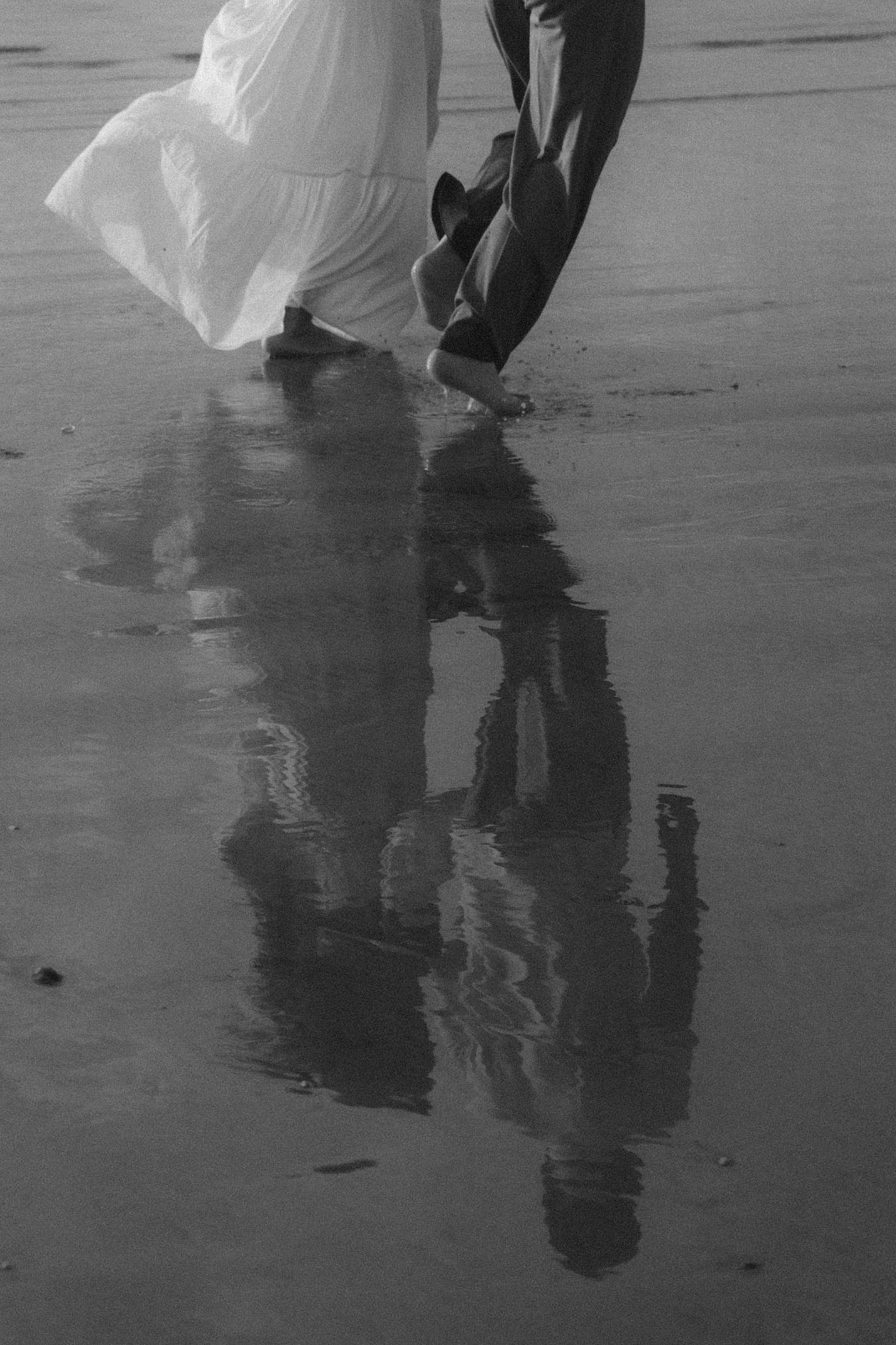 Black and white reflection of people walking on wet sand at the beach with flowing white dress creating mirror effect.