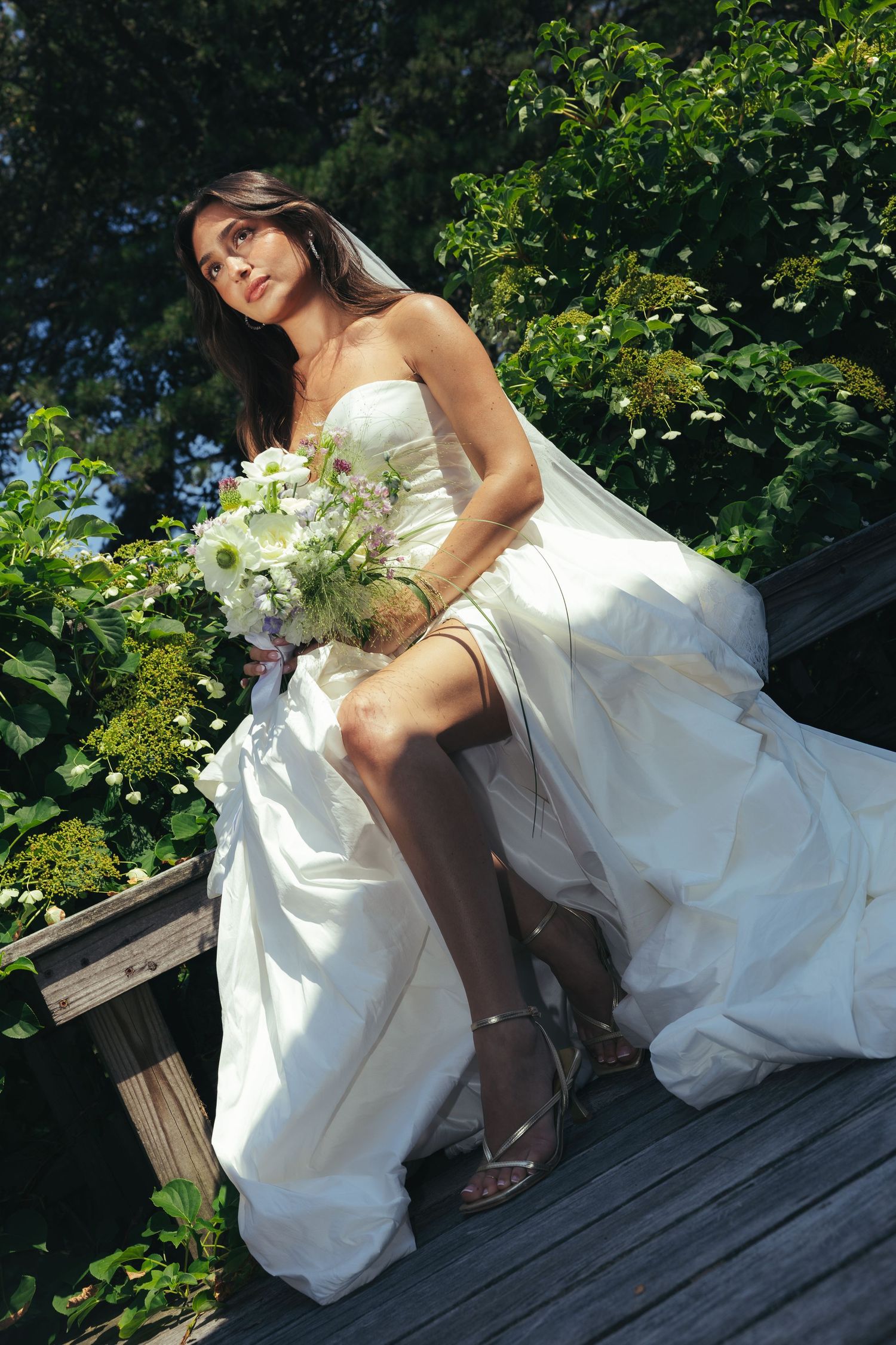 Bride in strapless white gown sitting on wooden deck holding floral bouquet surrounded by greenery.