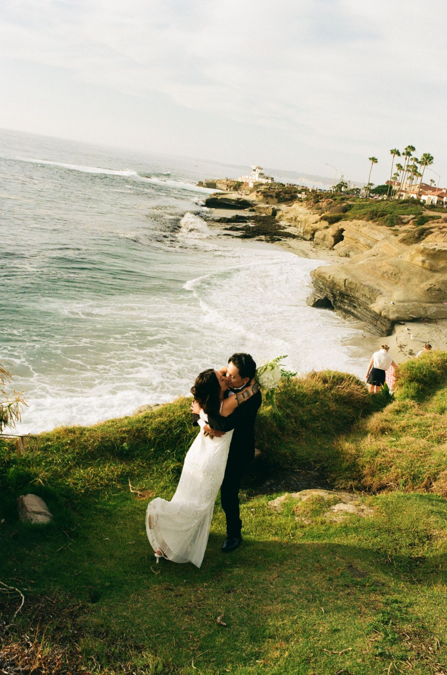 Bride and groom share a romantic kiss on a coastal cliff overlooking the ocean at their seaside wedding ceremony.