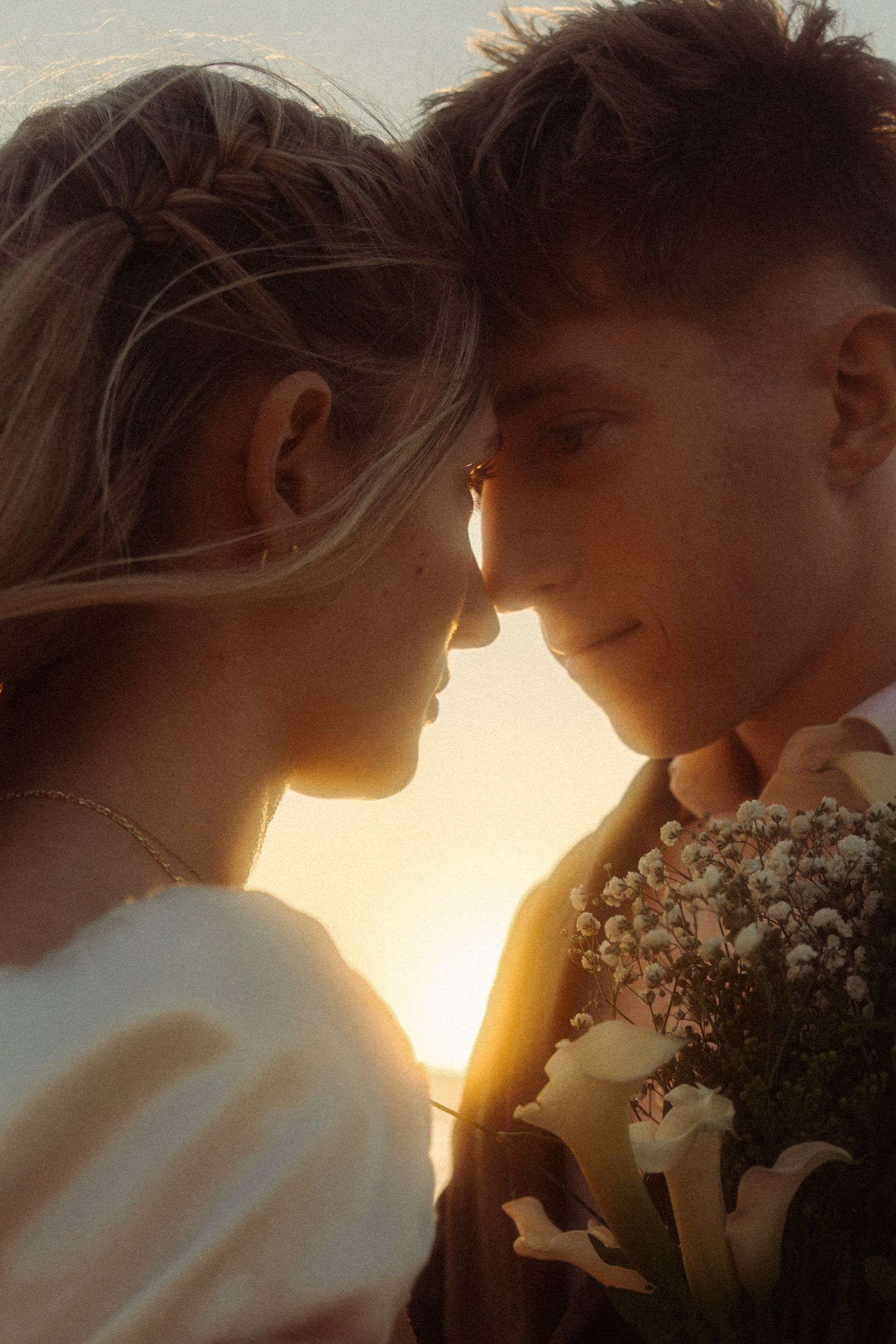 A couple shares a romantic sunset moment, foreheads touching, holding a bouquet of white flowers in golden light.