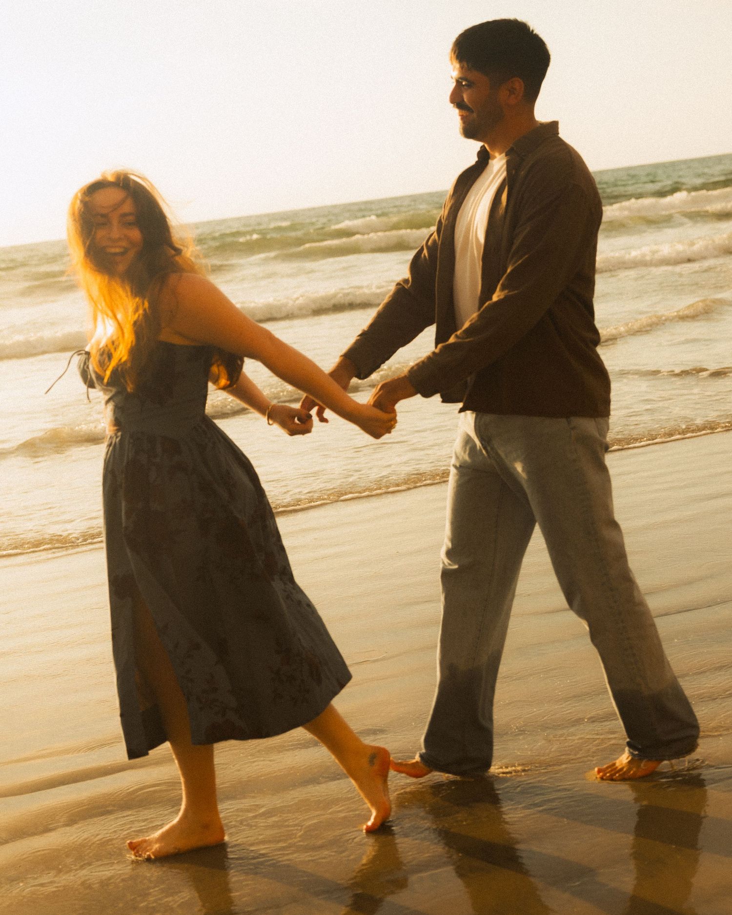 A couple dances playfully together on a sandy beach at sunset with waves in the background.