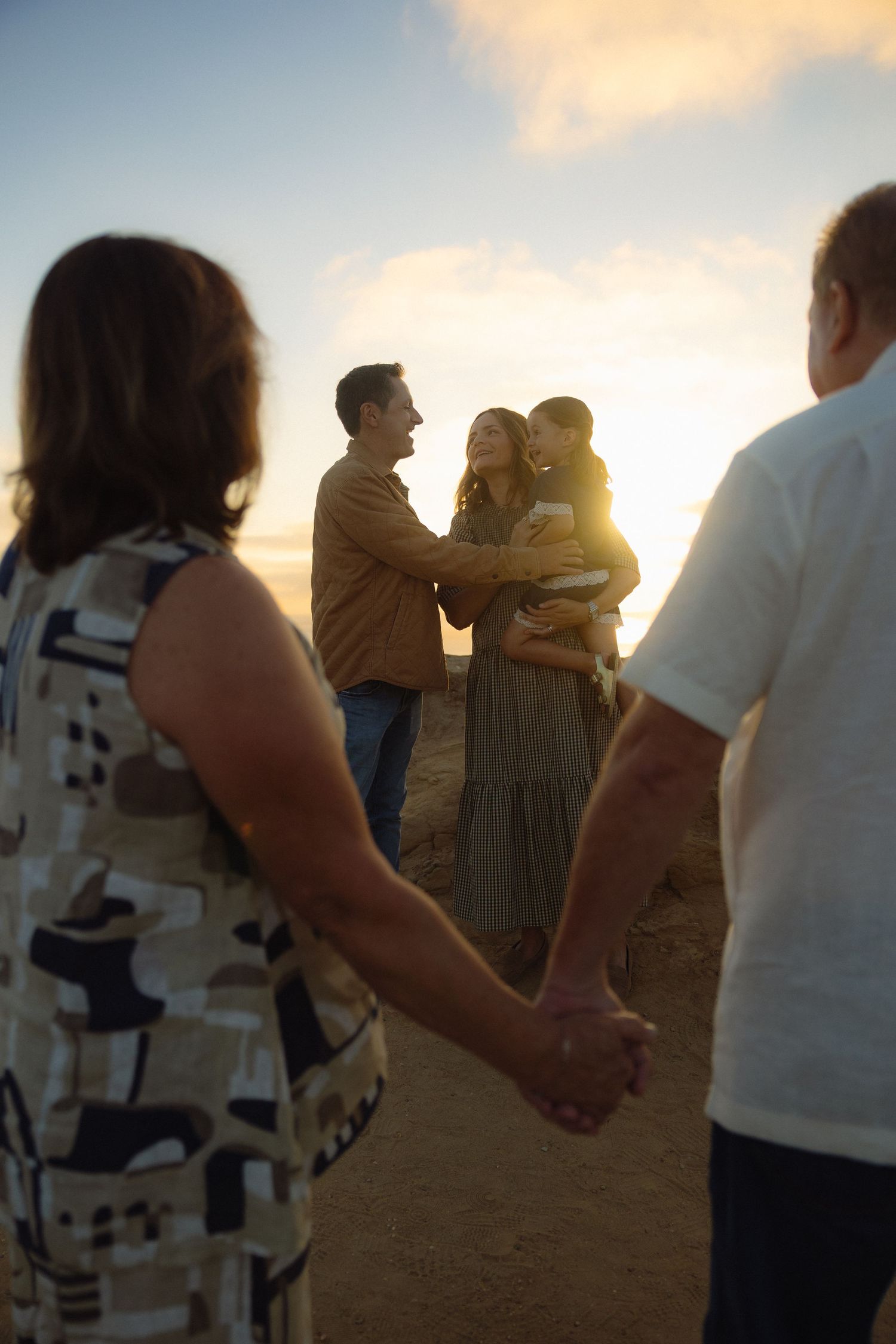 A group of people holding hands in a circle at sunset on a beach, creating a silhouette against the golden sky.