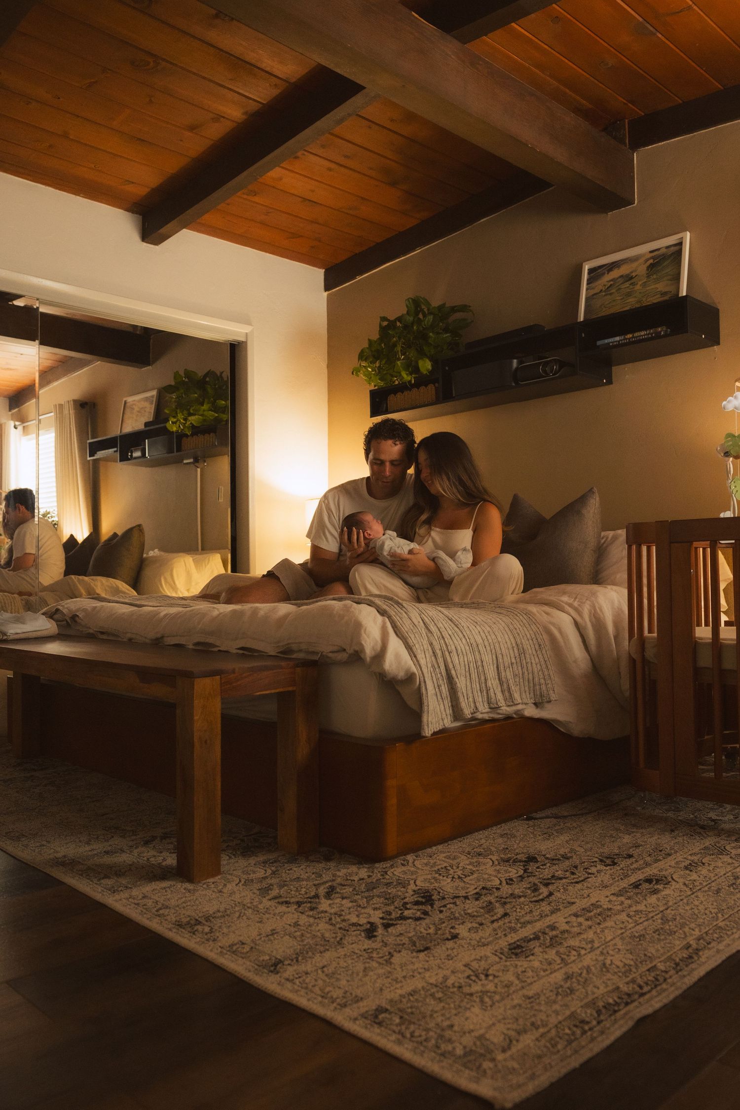 Cozy bedroom interior with wooden ceiling beams, warm lighting, and a comfortable bed with throw blankets.