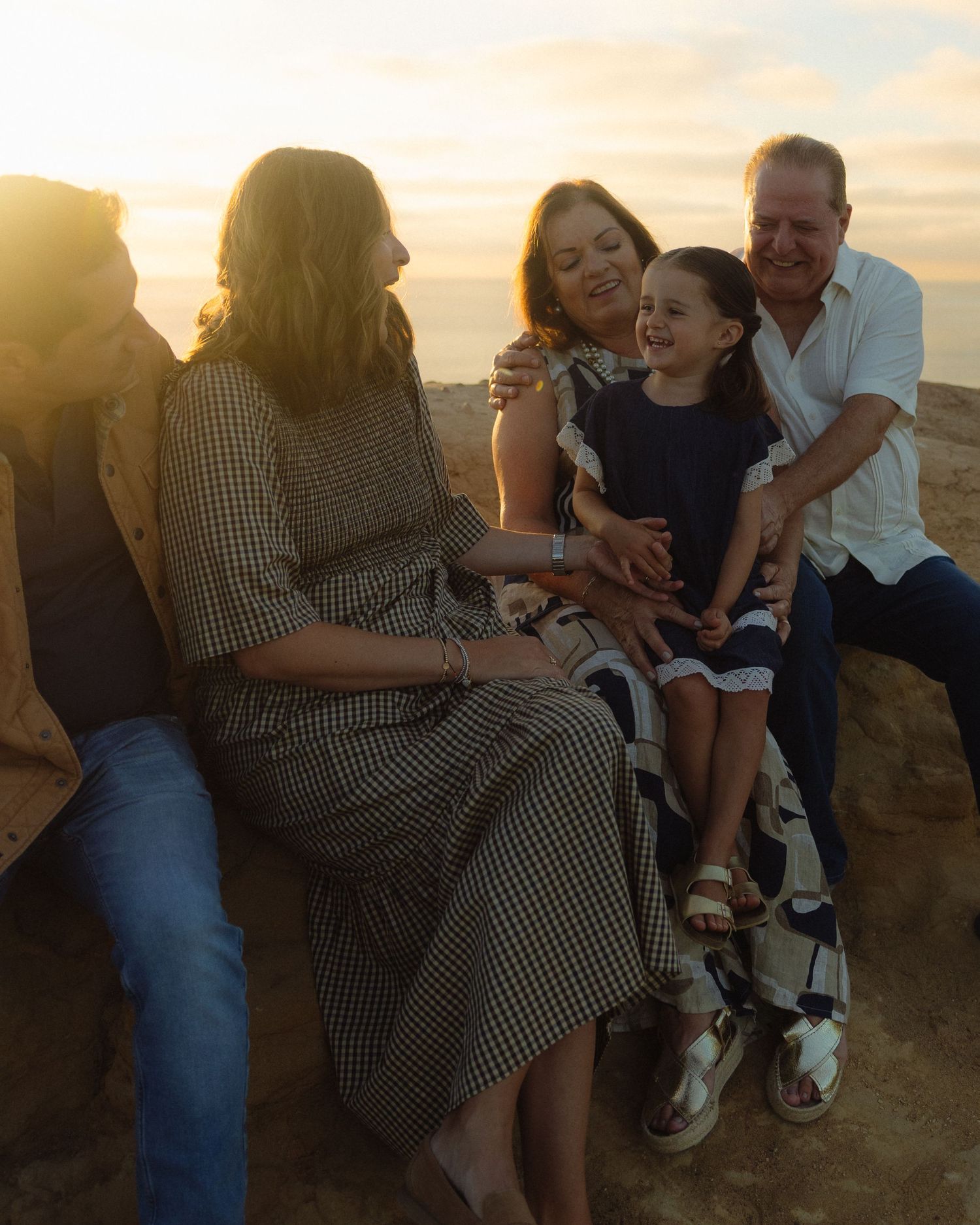 Family sits together on rocks at sunset watching ocean waves as golden light illuminates the peaceful coastal scene.