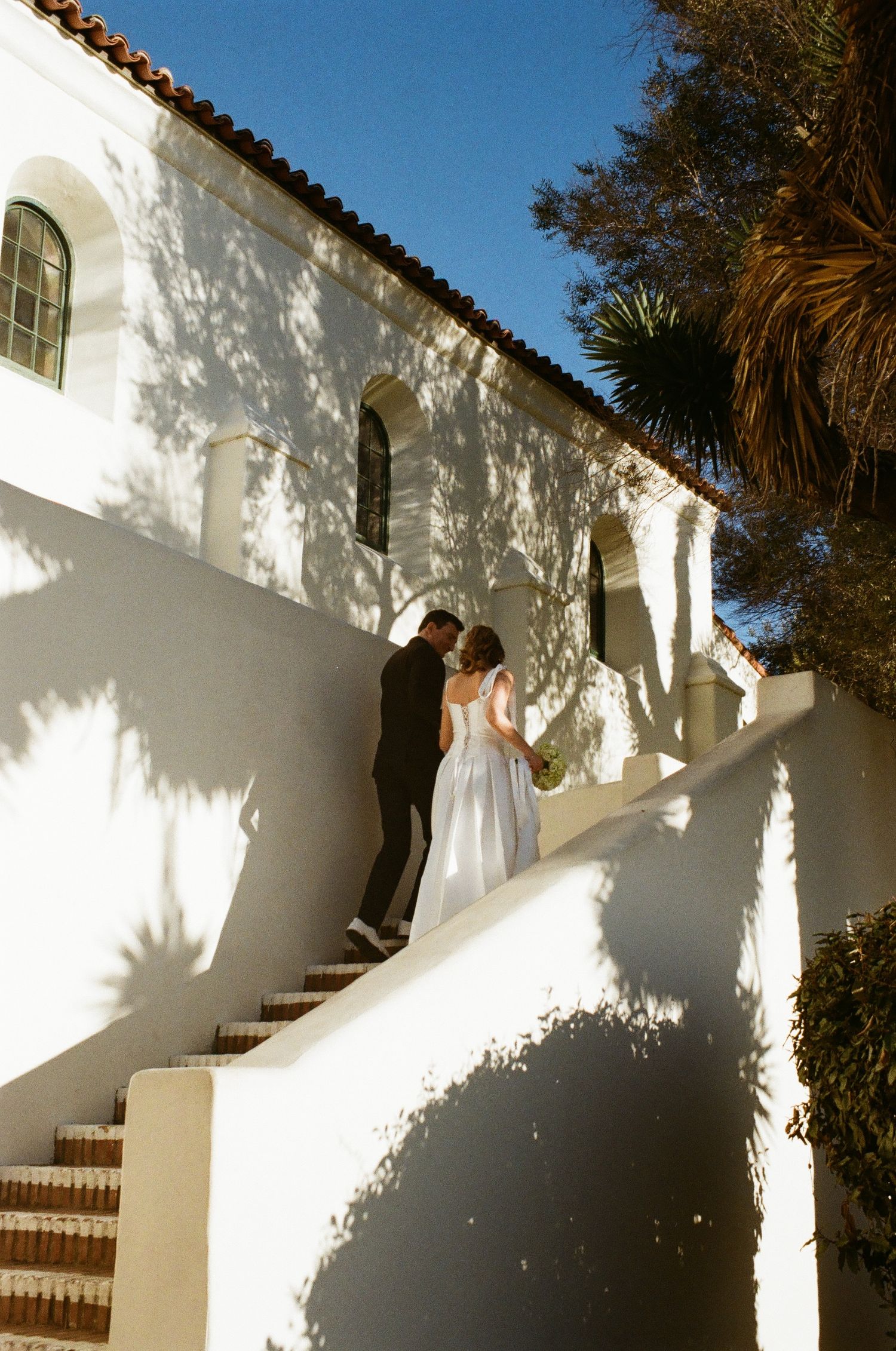 Person in white dress ascends curved staircase along Mediterranean-style white stucco building with terracotta tiles.