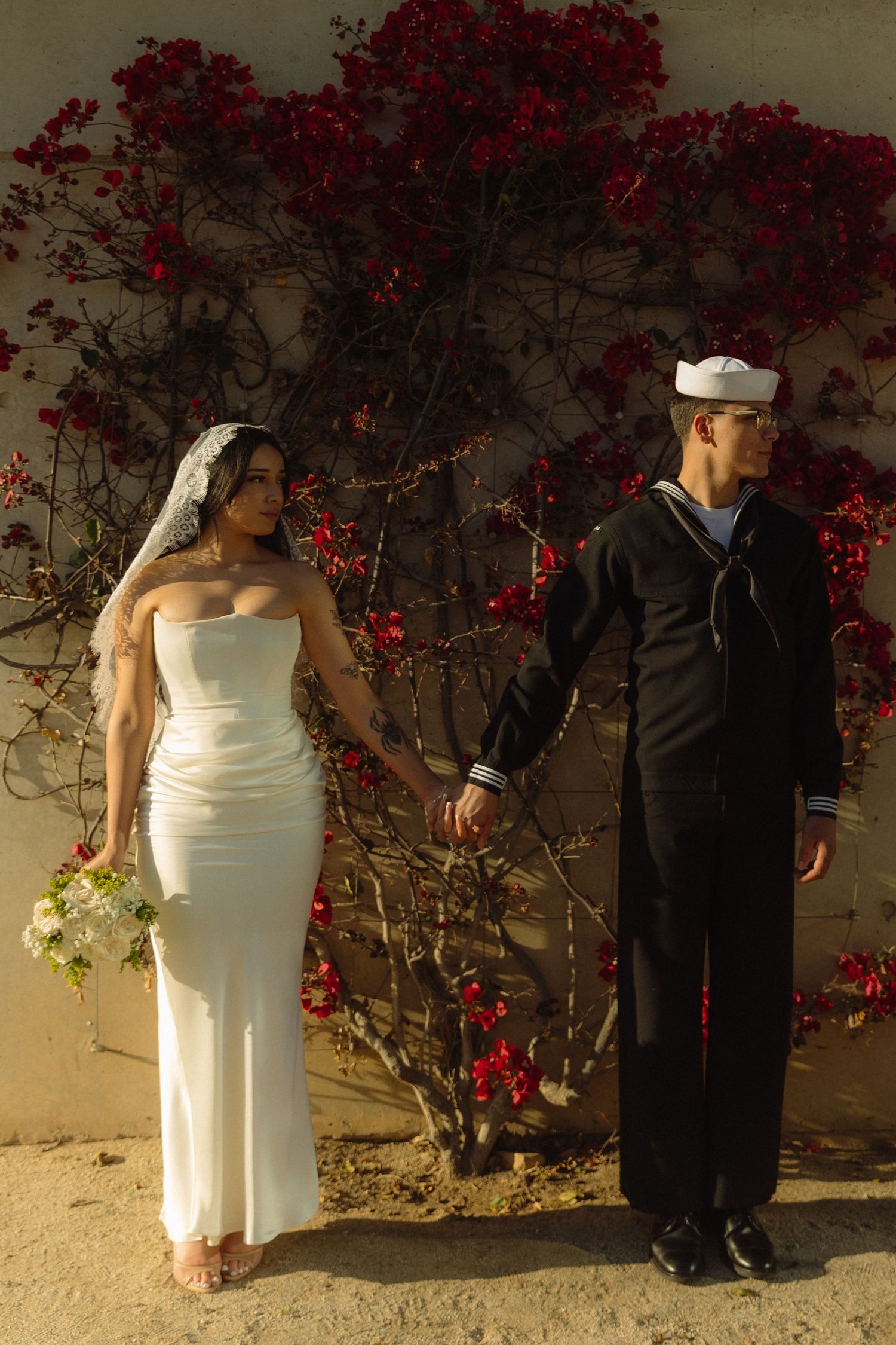 Navy sailor and bride hold hands in front of vibrant red flowering wall during outdoor wedding ceremony.