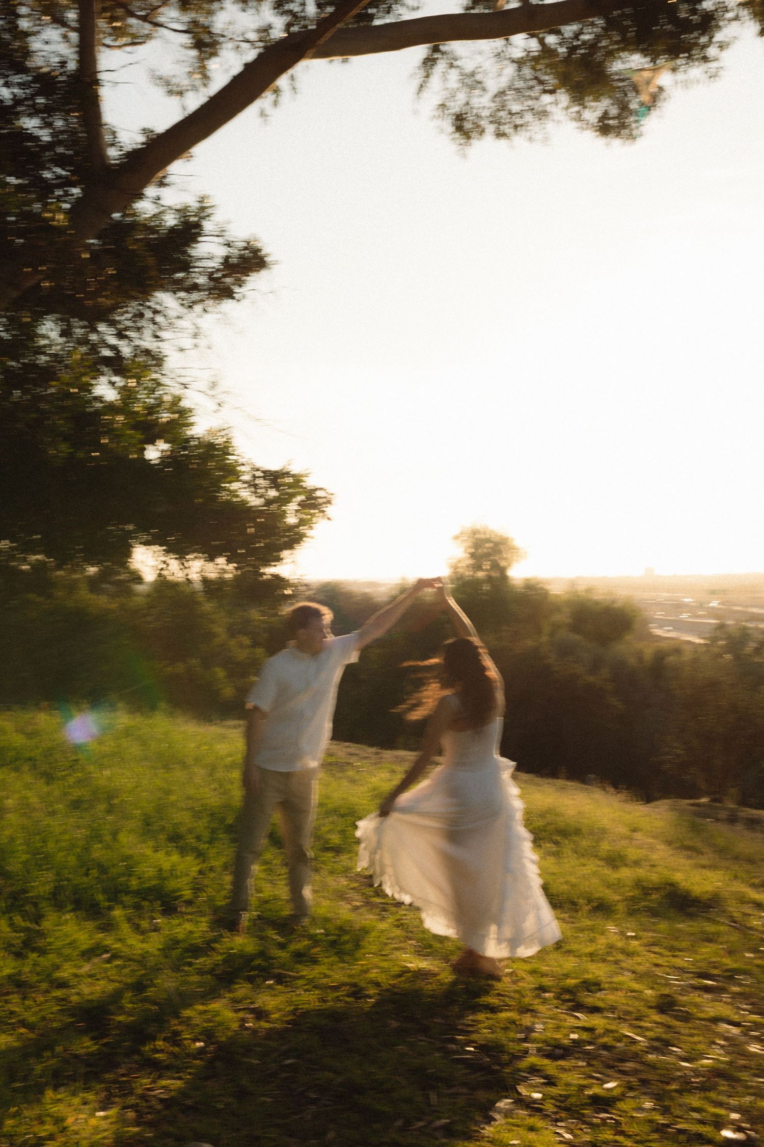 Couple dancing in golden sunset light on a grassy hill, woman in white dress twirling under trees.