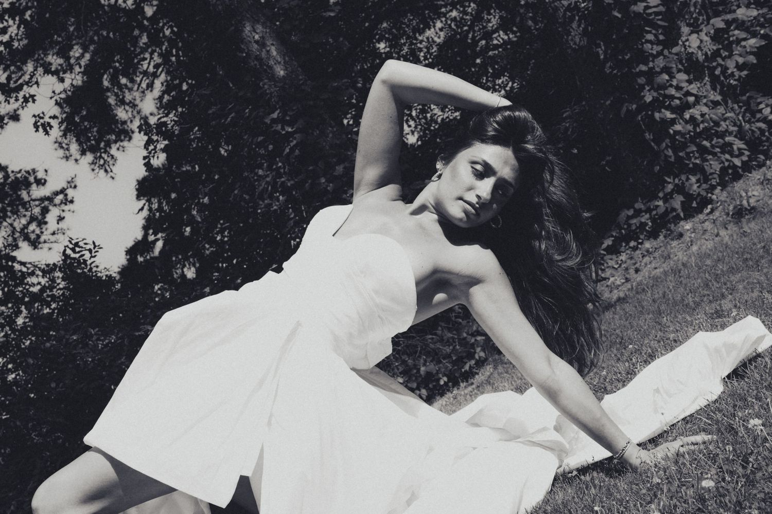 Black and white photo of bride in white dress posing dramatically on grass near trees.