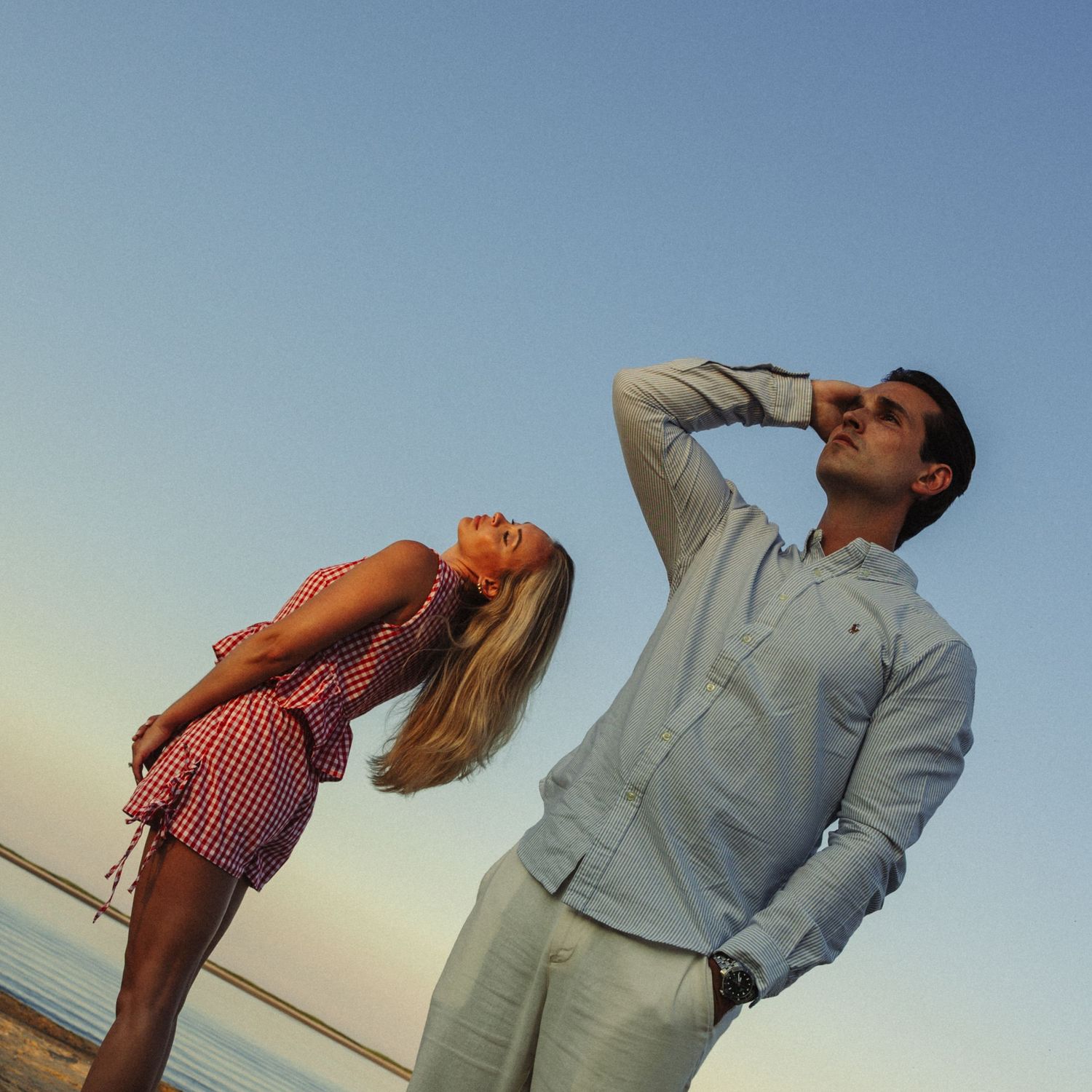 A couple poses at sunset on a beach, woman in red dress leaning back, man in light blue shirt looking away.